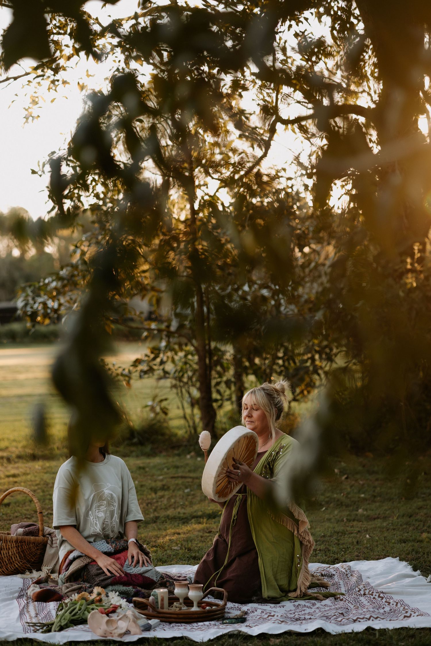 Two women enjoying a golden hour picnic on a blanket under a large tree in a lush green field.