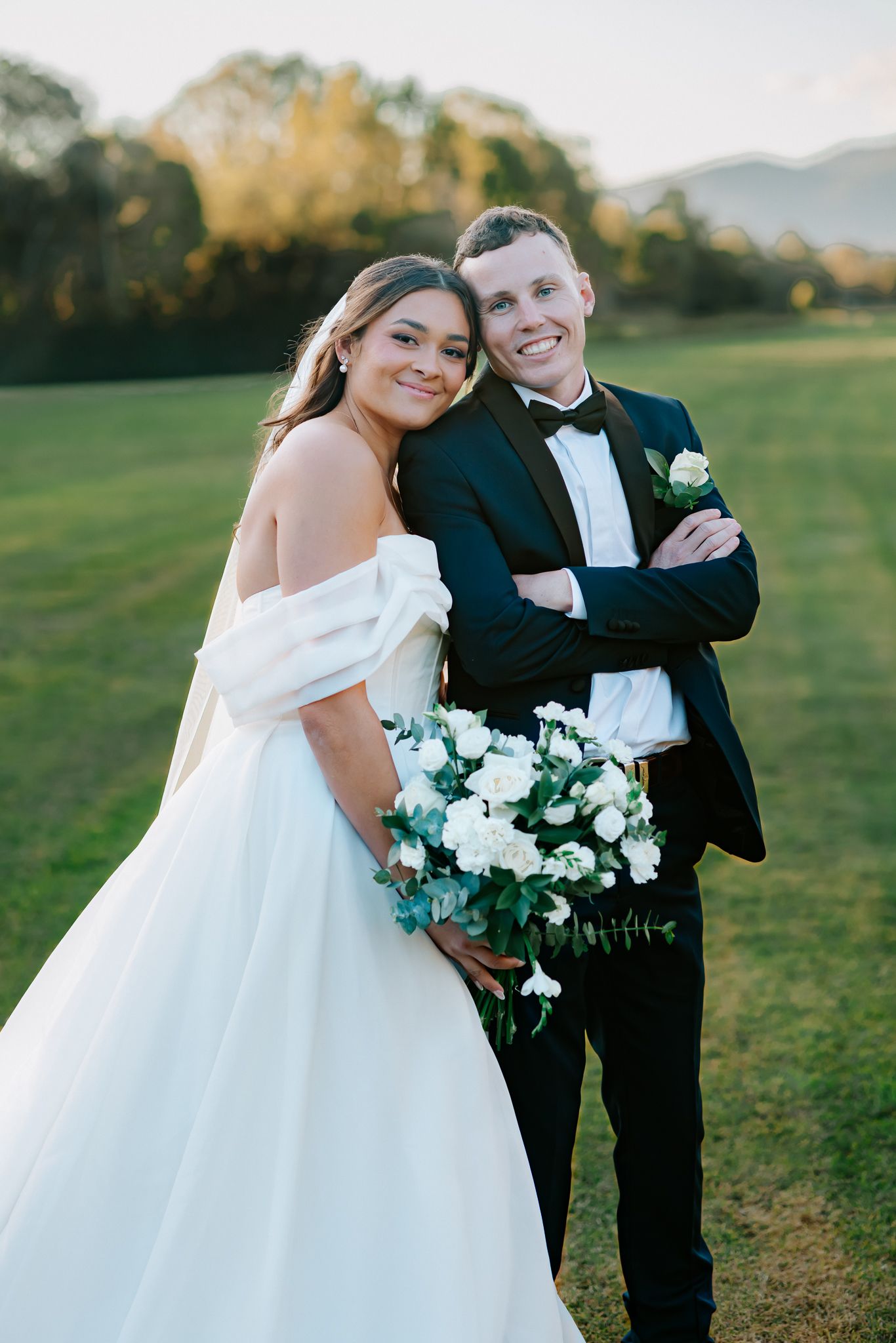 Smiling bride in off-shoulder gown leans on groom in black tuxedo holding white bouquet outdoors.