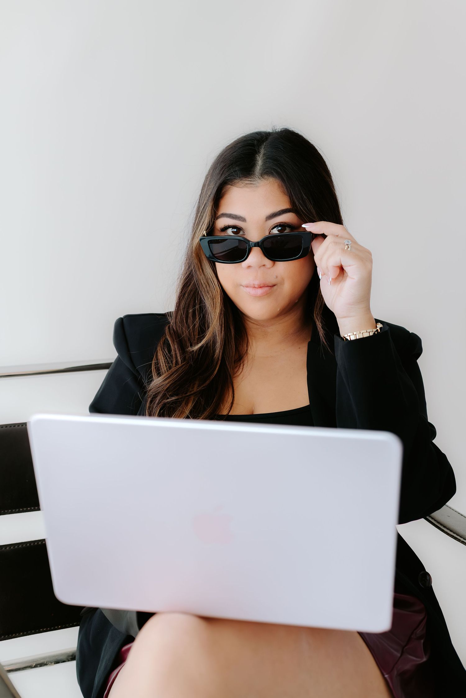 Young woman in black blazer and sunglasses working on laptop, peering over frames with confident expression.