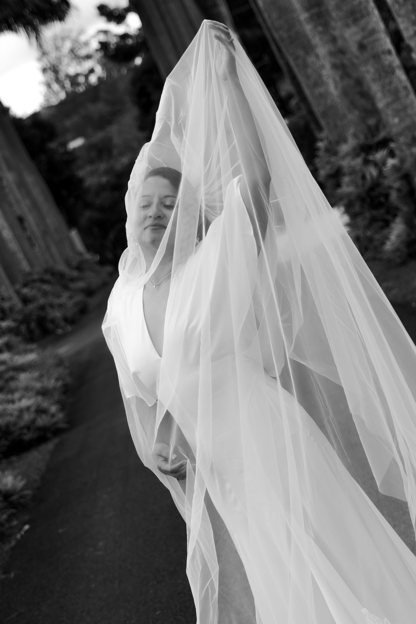 Black and white photo of a bride wrapped in a flowing wedding veil outdoors on a garden path.