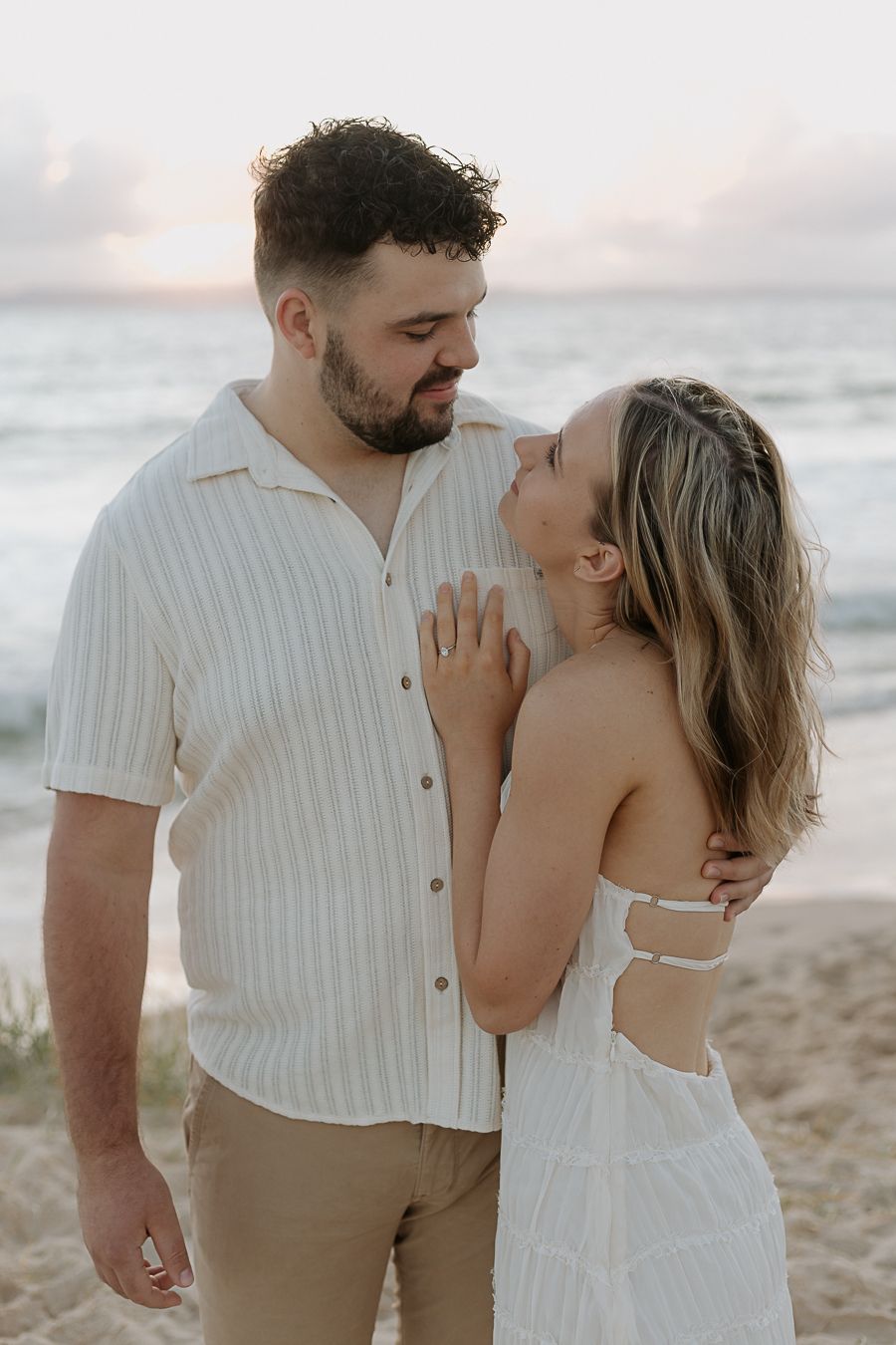 Engaged couple sharing a tender moment on the beach, woman in white sundress touching mans striped shirt.