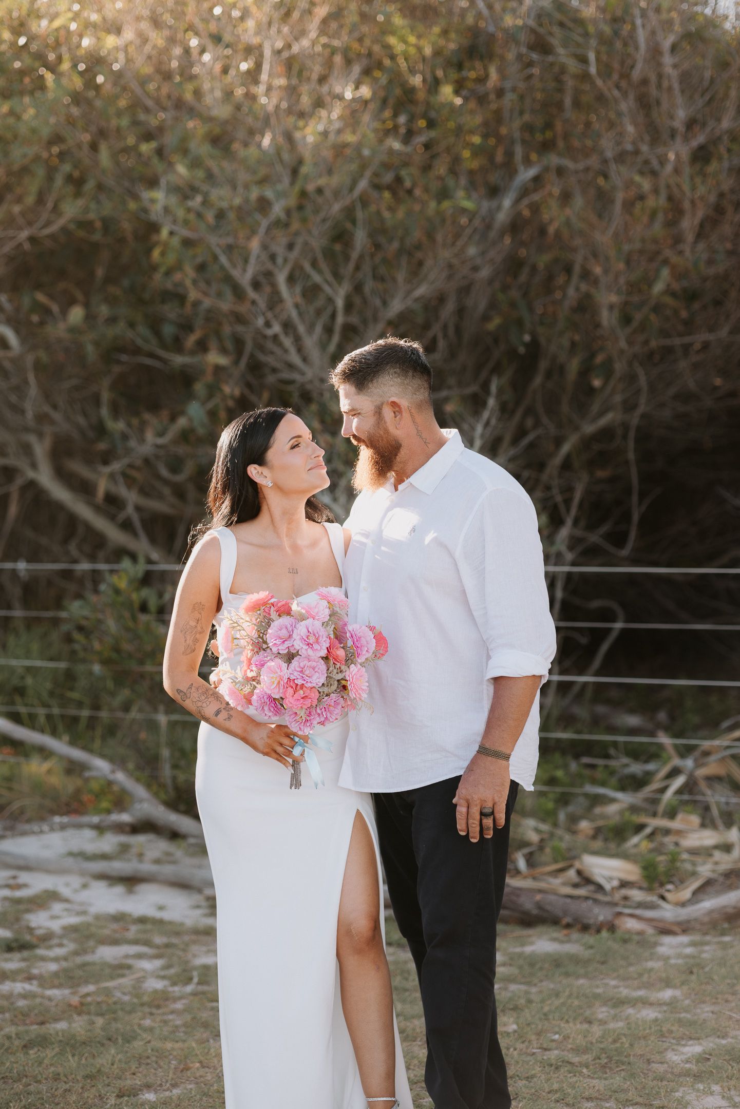 A couple in wedding attire shares romantic moments outdoors with pink floral bouquet during their photoshoot.