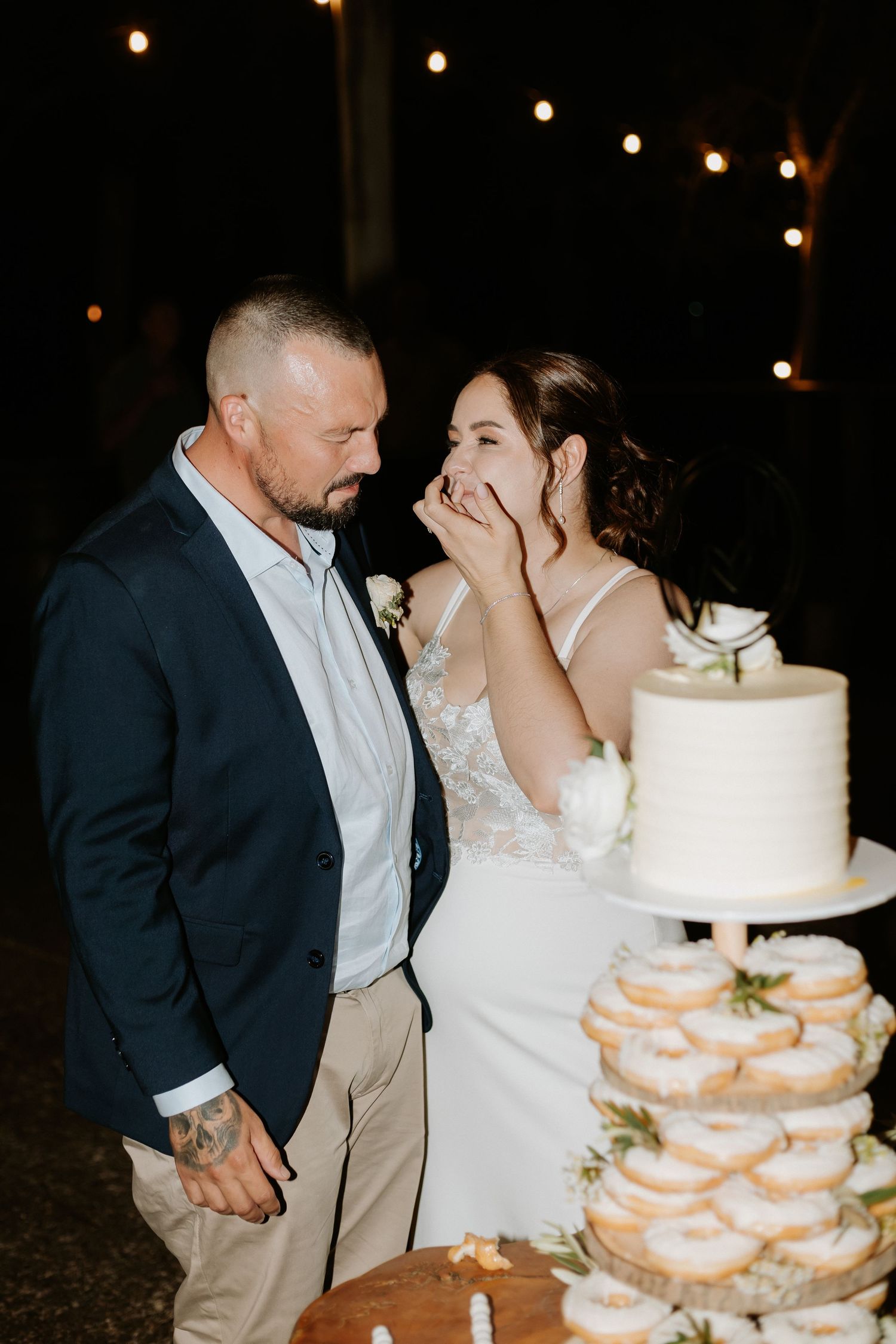 Wedding couple sharing a moment by their tiered white cake with string lights in background.