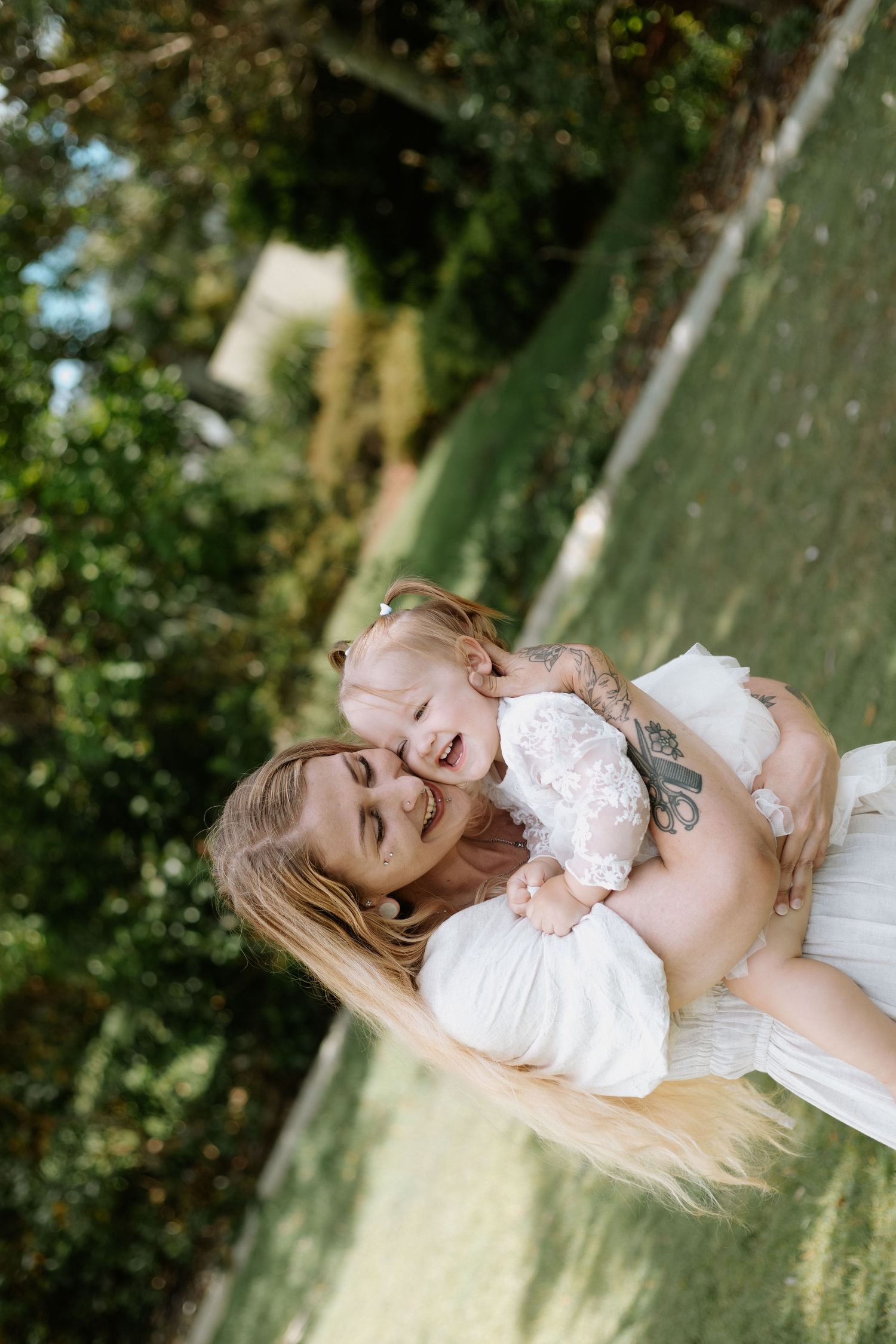 A parent and baby wearing white outfits share a joyful moment together on grass in an outdoor setting.