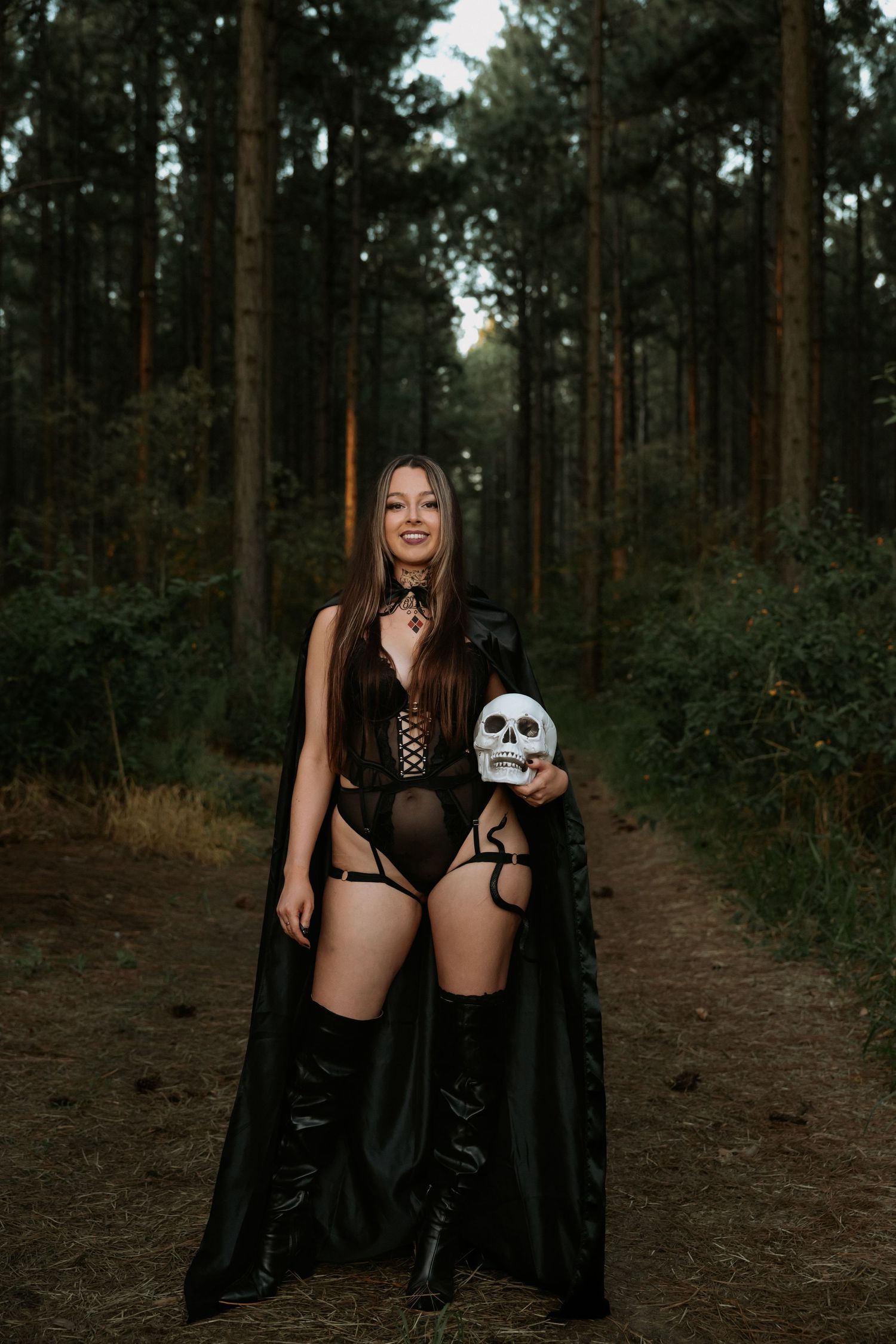 Woman in black Halloween costume holding skull prop in forest setting.