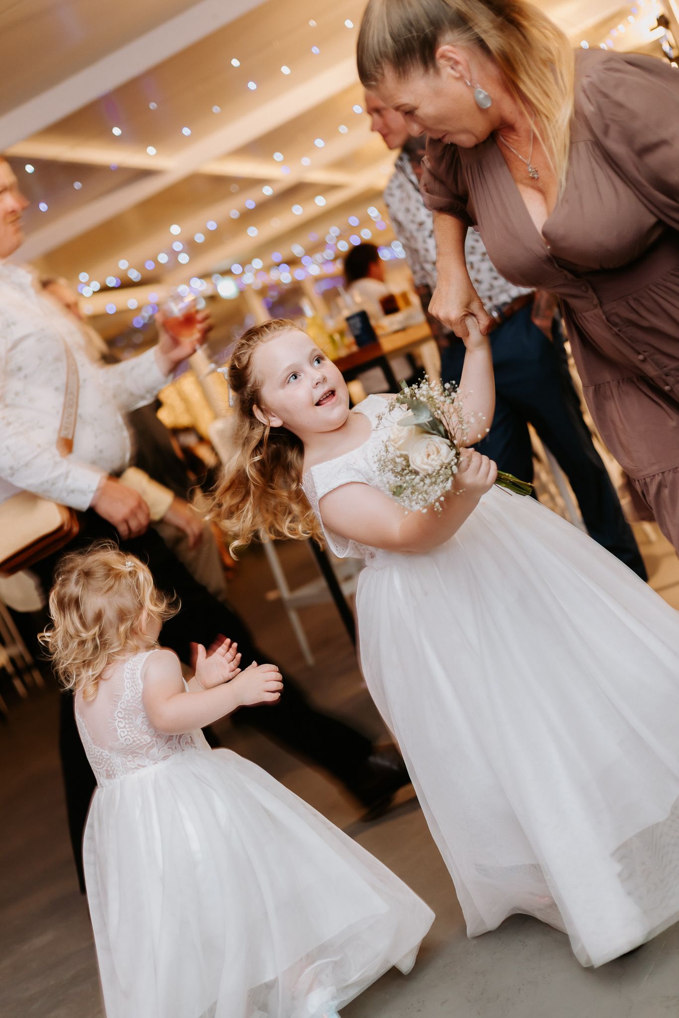 Two young flower girls in white dresses dancing joyfully at a wedding reception with guests nearby.