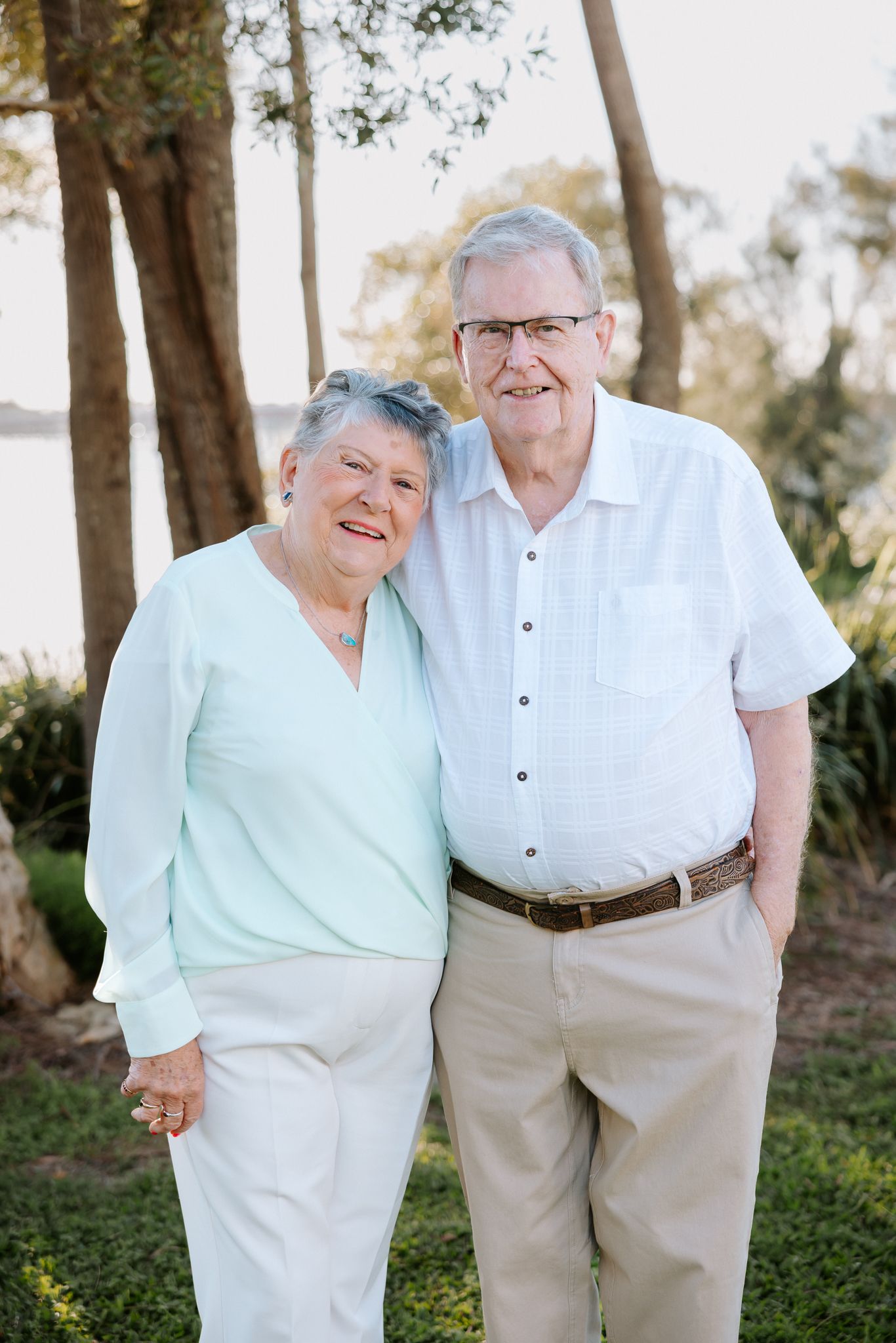Smiling elderly couple posing outdoors near trees and water, wearing light casual clothing on a sunny day.