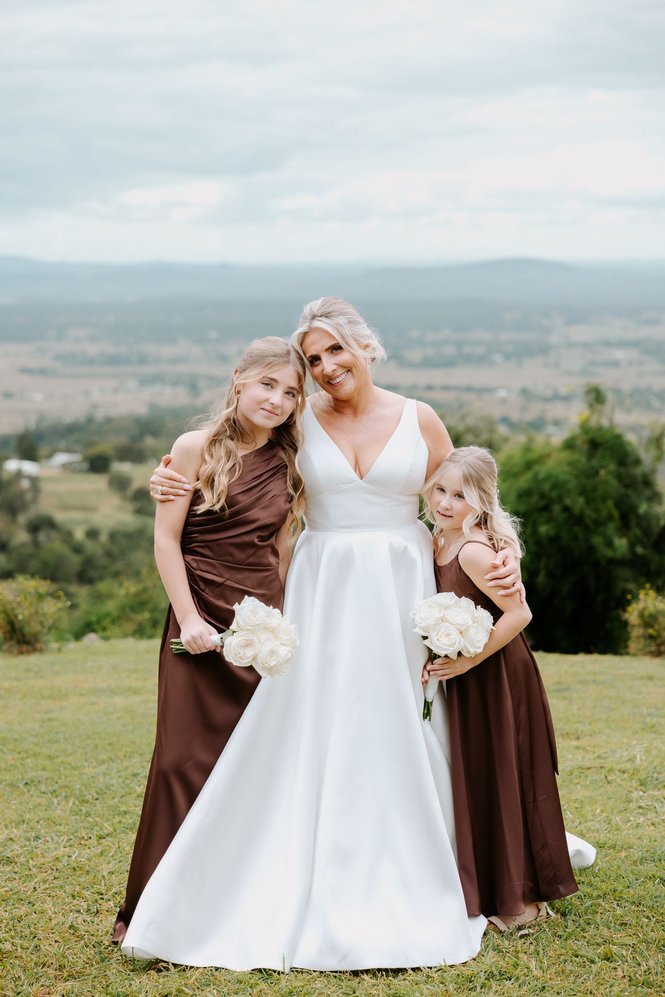 Bride in white gown poses with two bridesmaids in brown dresses holding white bouquets outdoors.