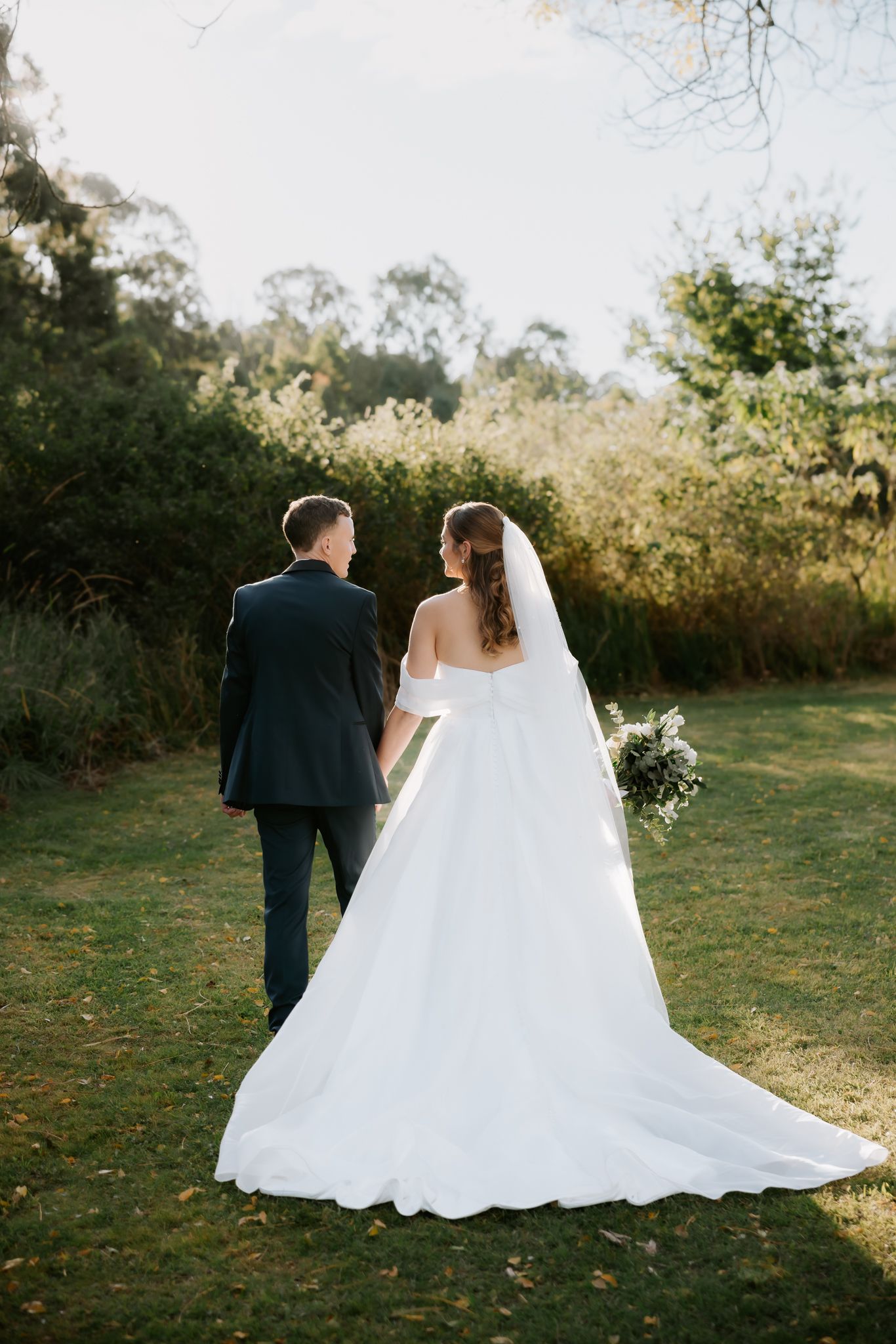 Bride and groom walking hand in hand through lush green garden, bride wearing elegant white gown with long train and veil.