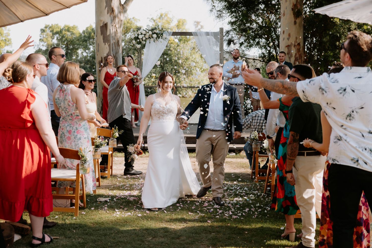 Outdoor wedding celebration with confetti toss under trees and white tent.