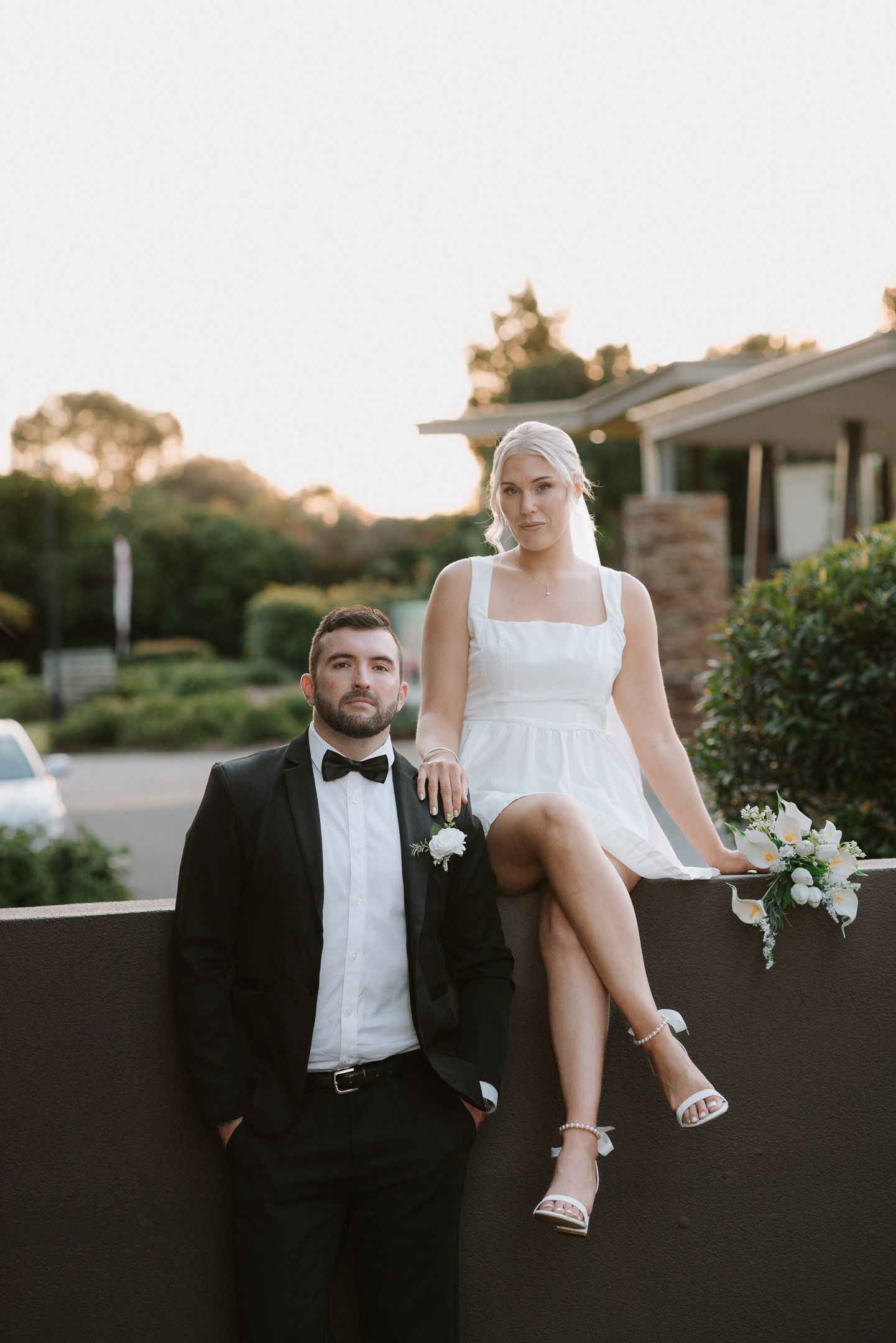A couple in formal attire posing casually outdoors at sunset.