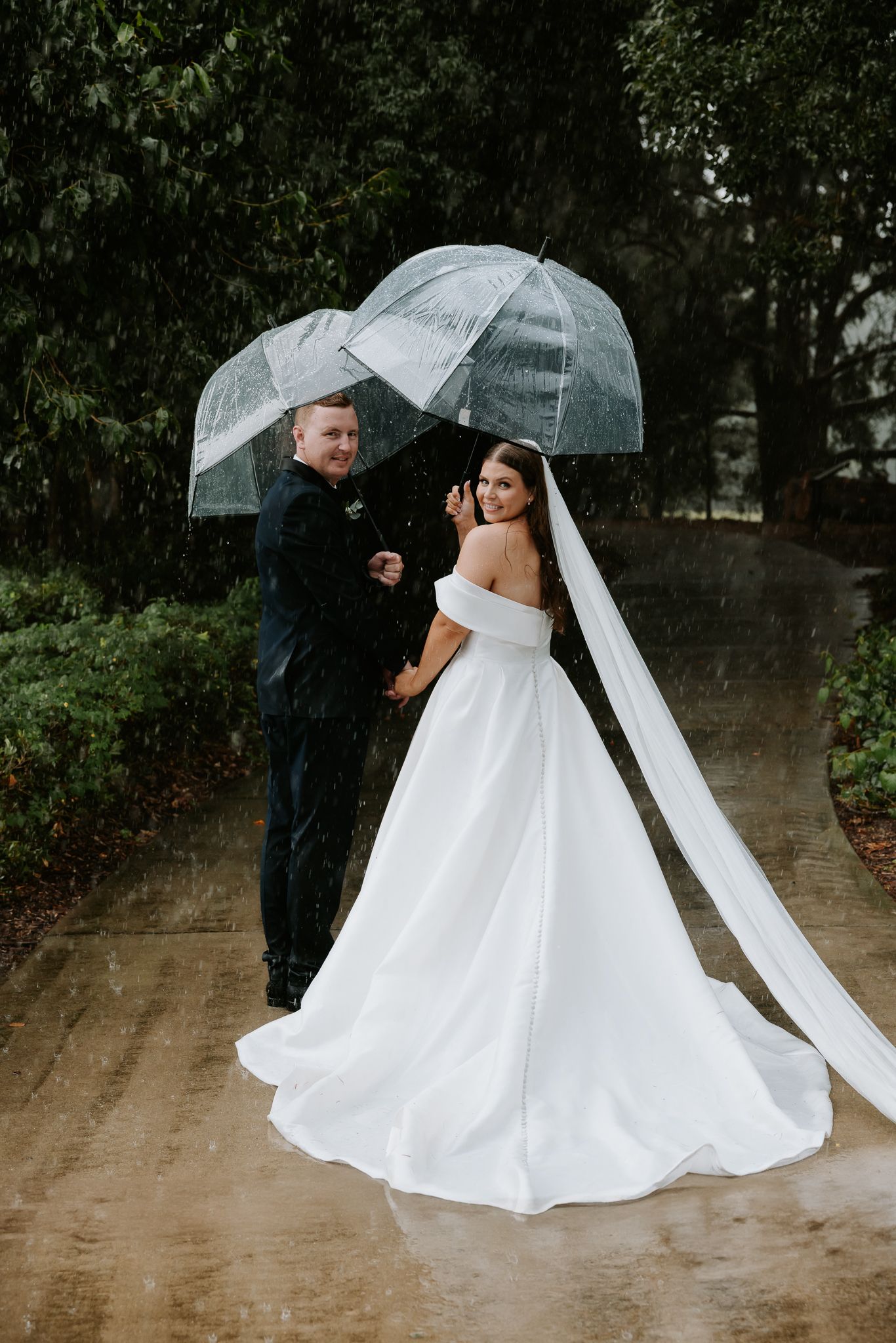 A couple shares clear umbrellas in the rain on their wedding day, with the bride wearing an elegant off-shoulder white gown.