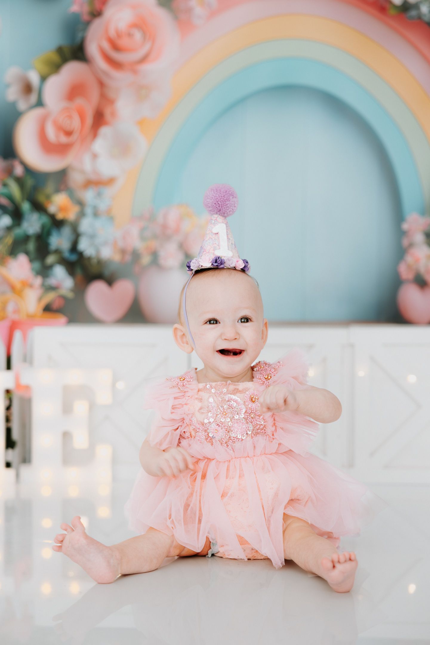 A baby in a pink ruffled dress sits in front of a pastel rainbow backdrop with roses and decorative elements.