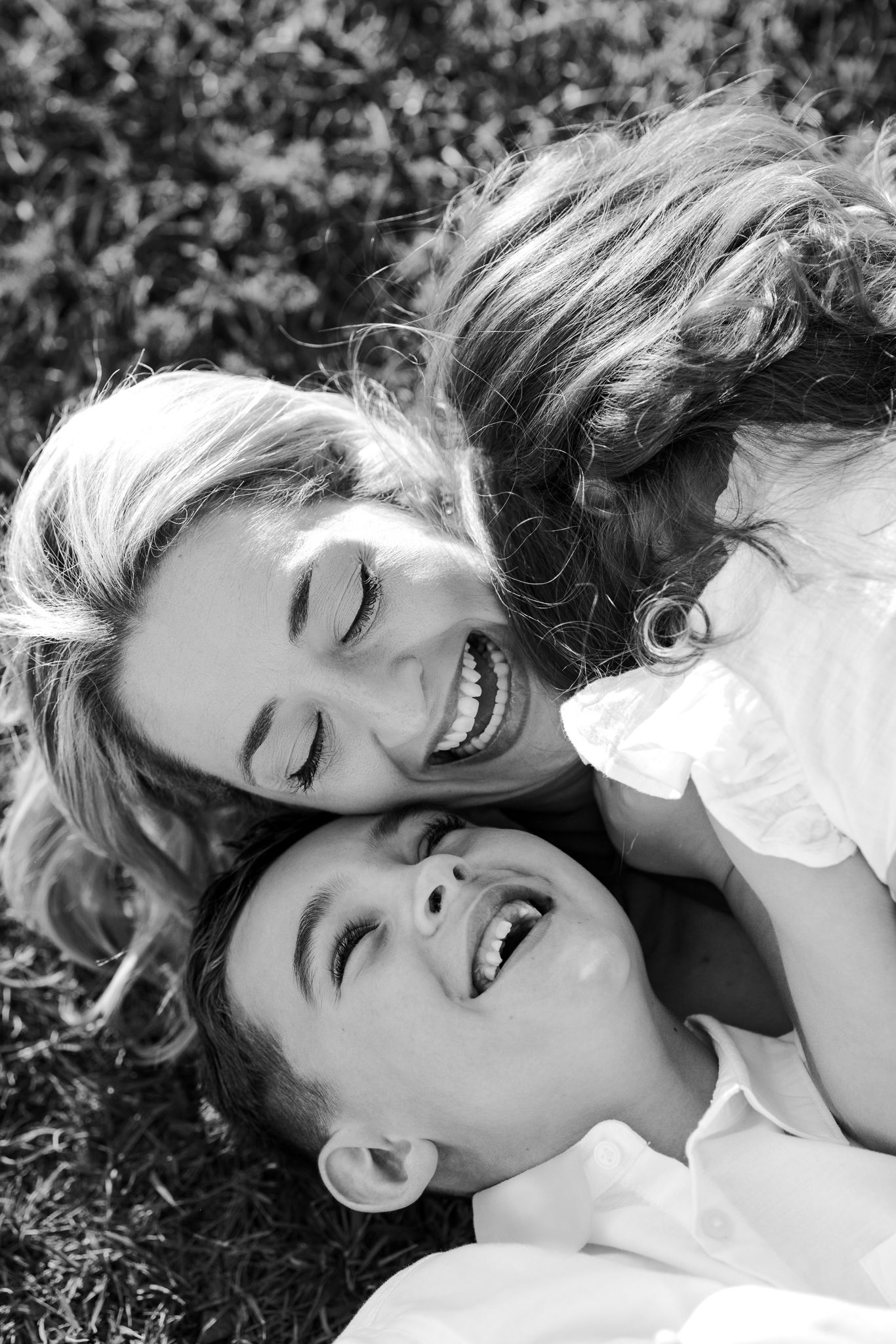 Black and white photo of a mother and two children laughing together while lying on grass outdoors.
