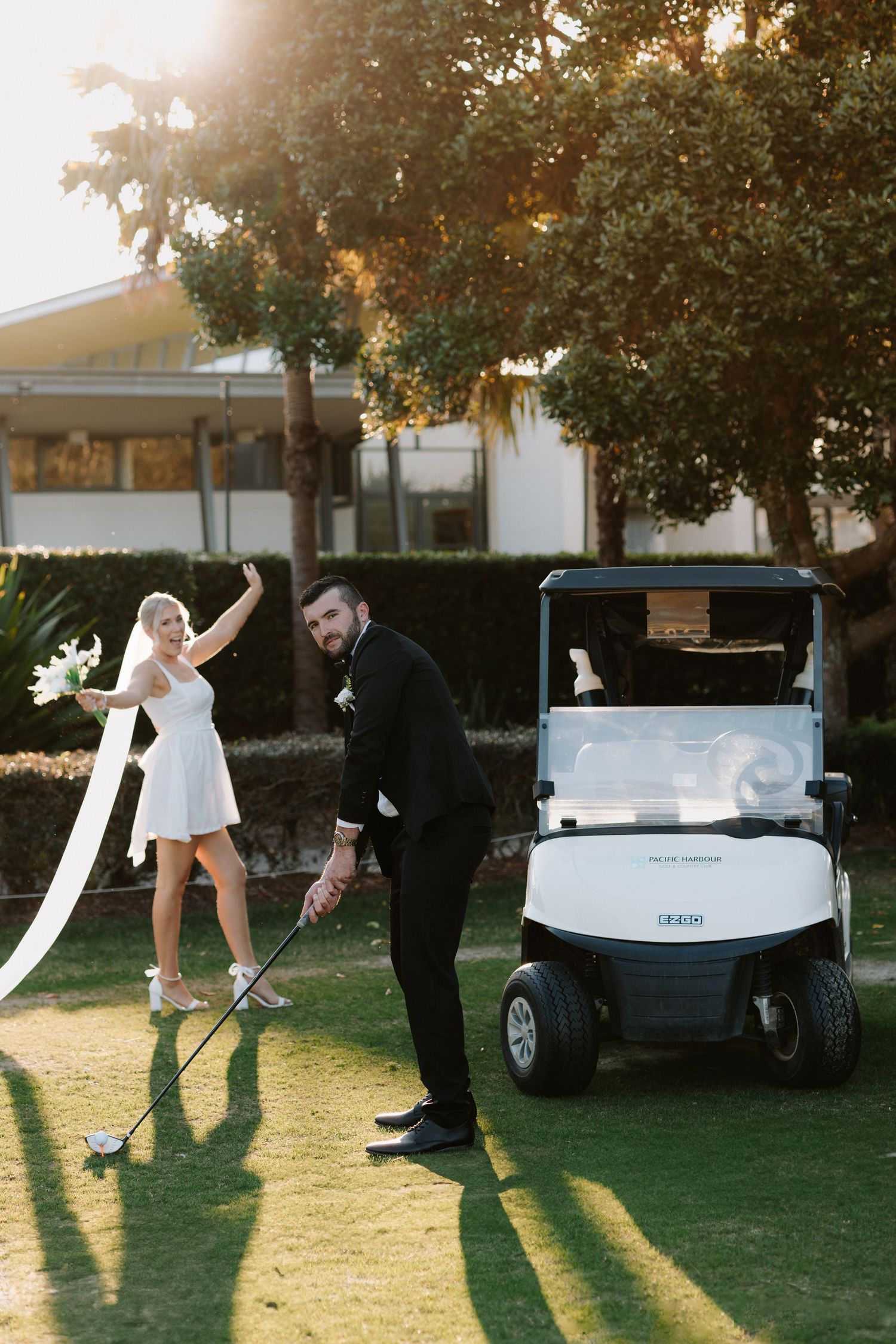Bride and groom posing on golf course near white golf cart, bride waving bouquet in short wedding dress.