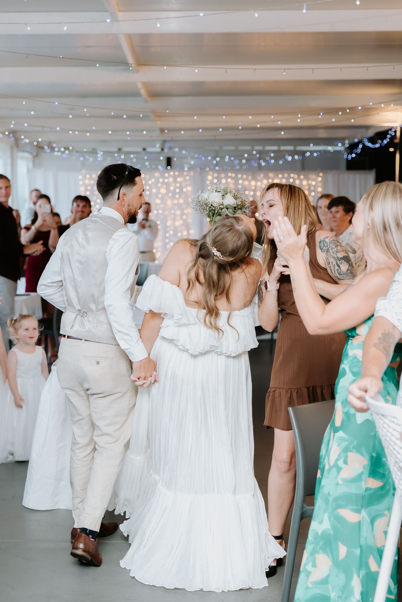 Bride and groom entering wedding reception hall decorated with fairy lights, greeted by excited guests.