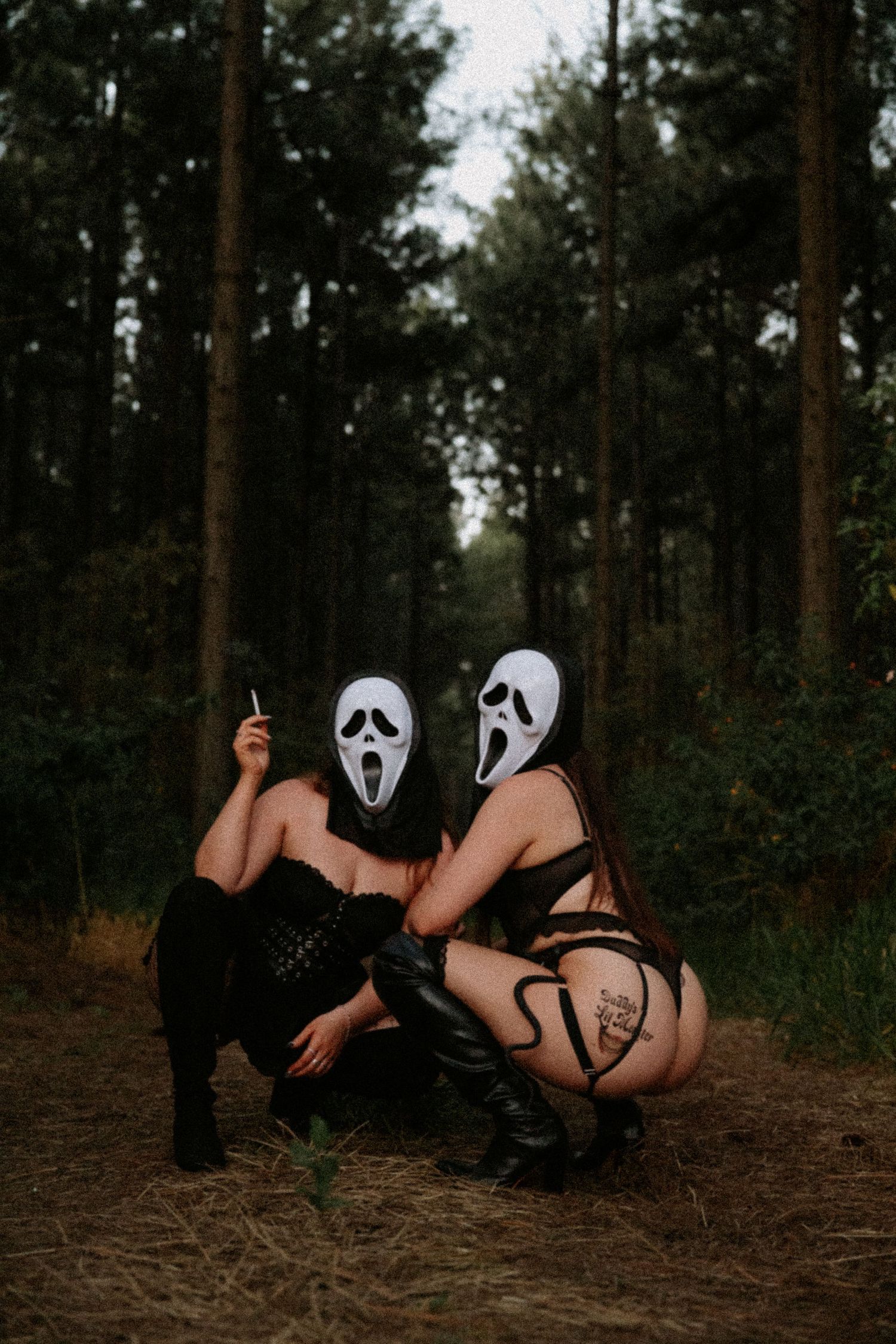 Two people wearing Ghostface masks crouch in a dark forest wearing Halloween costumes.