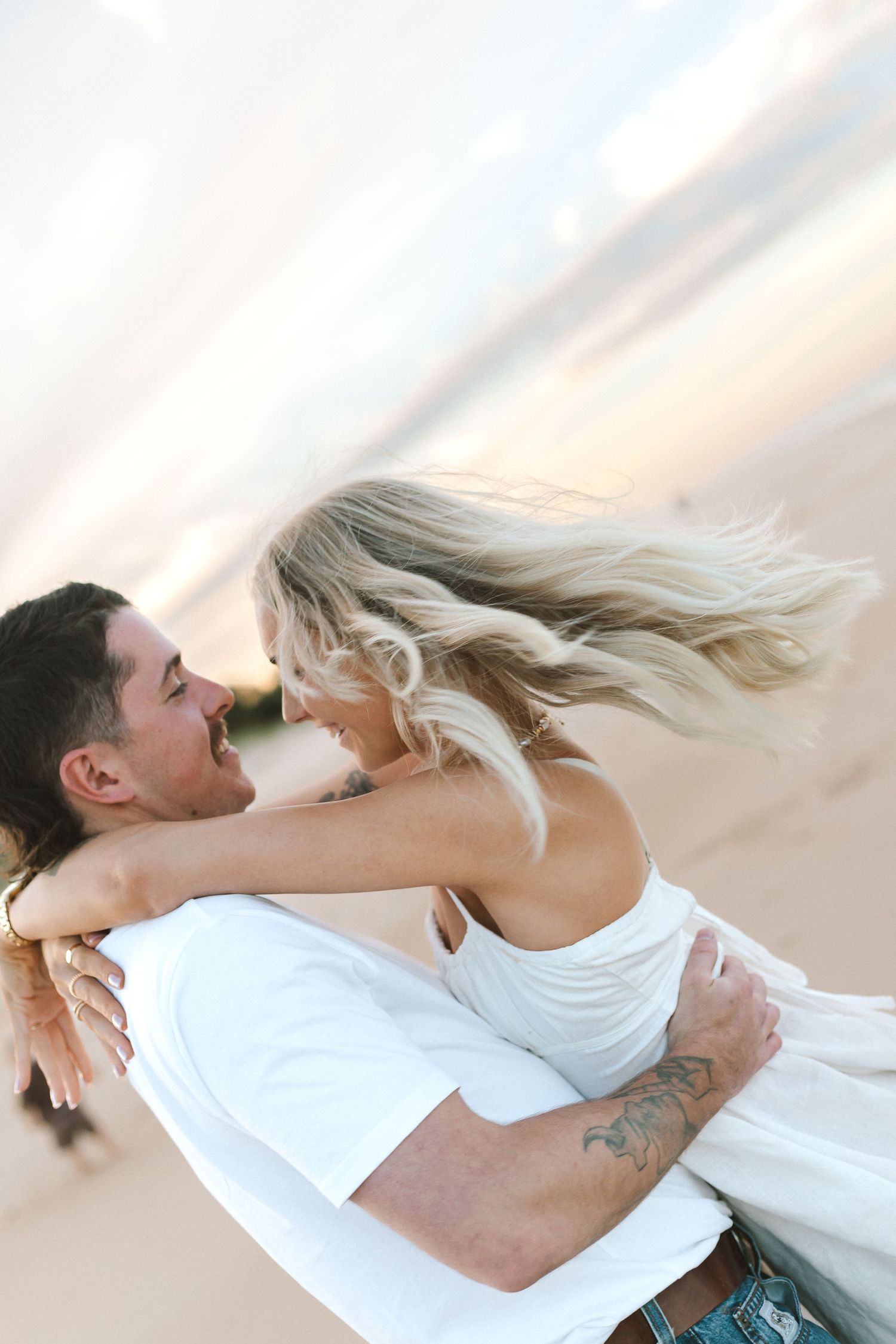 A joyful couple embraces on a sandy beach at sunset, the woman laughing as her blonde hair blows in the wind.