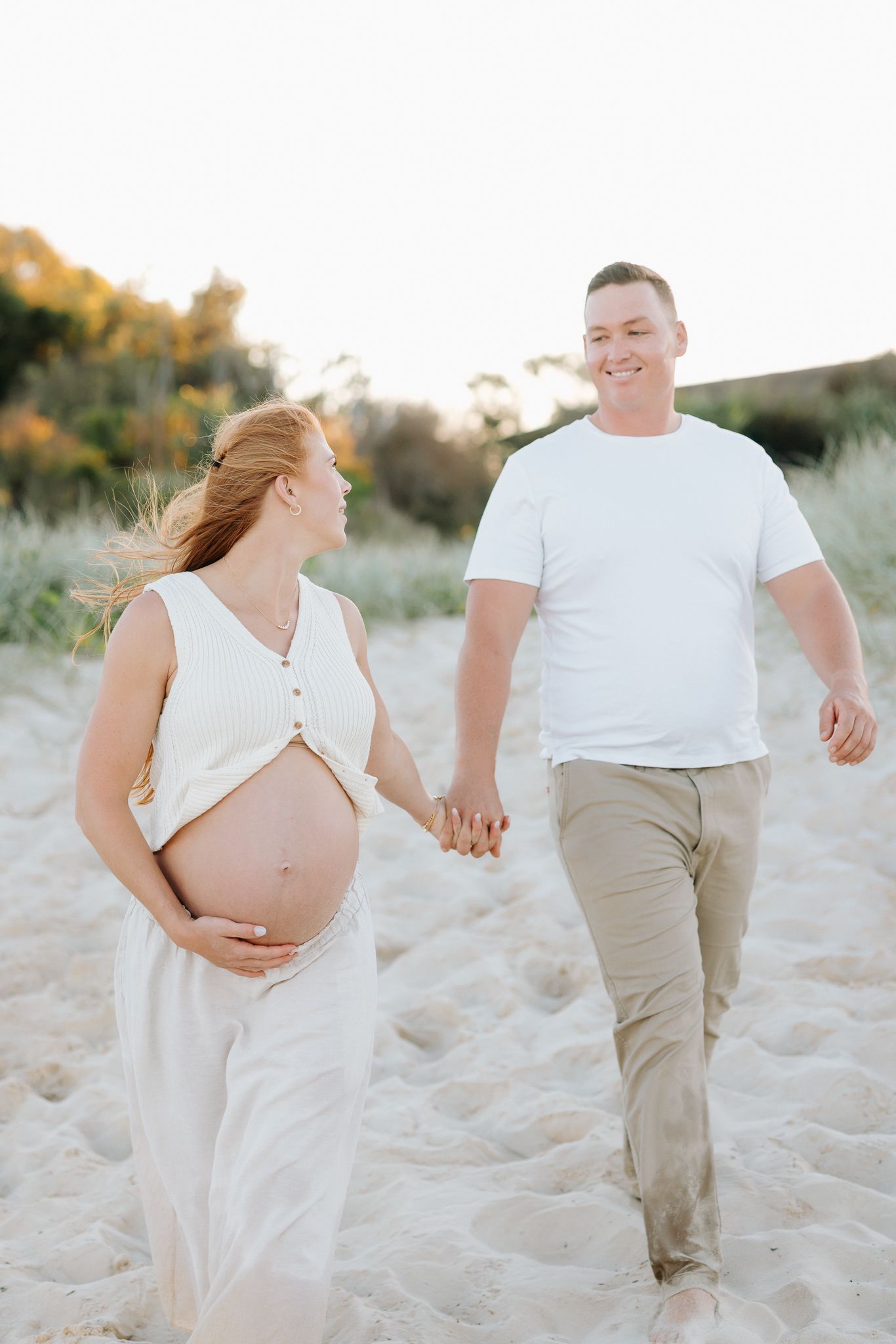 Pregnant couple holding hands walking on sandy beach, woman in white dress cradling baby bump, man in white tee and khaki pants.