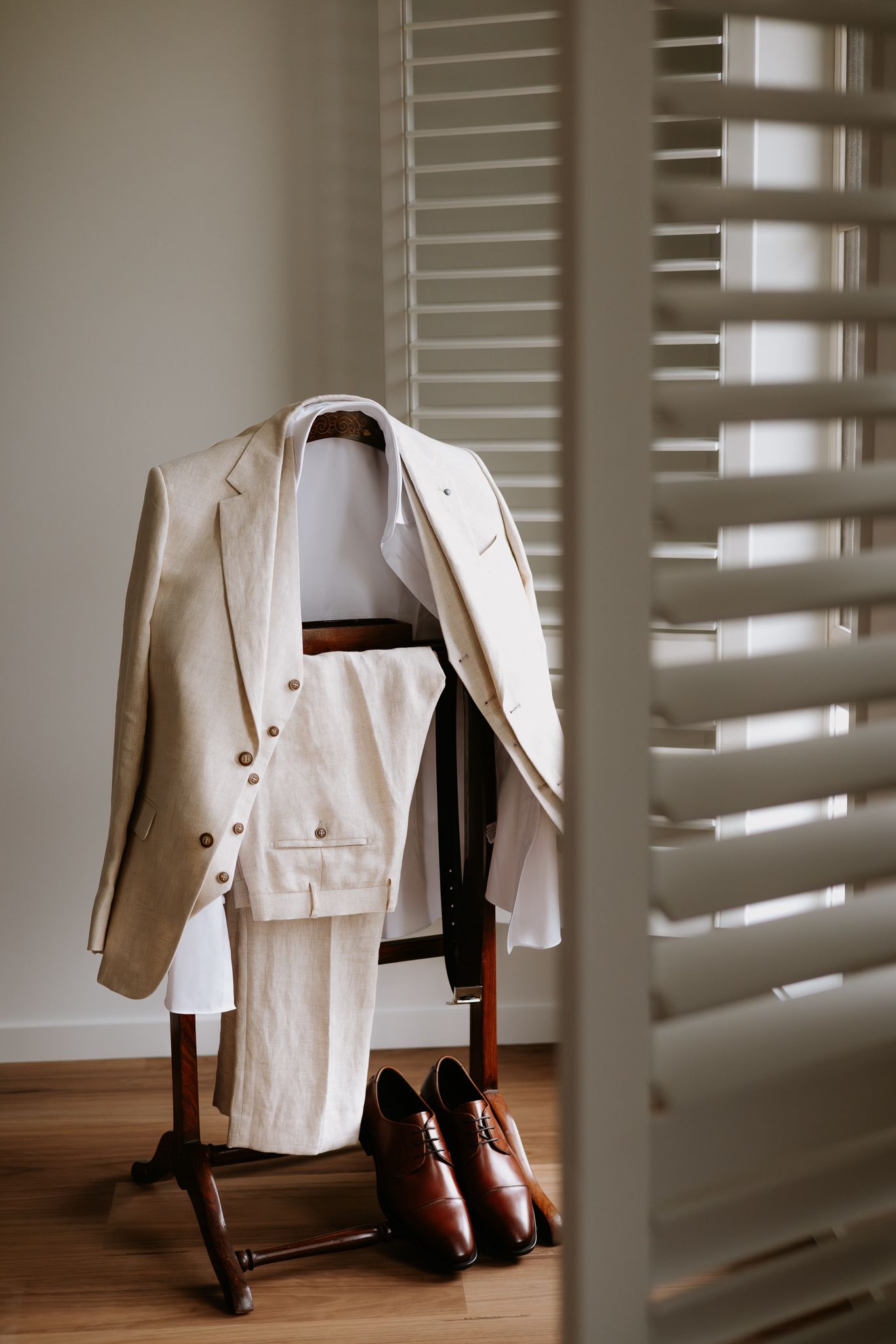 Beige linen suit on valet stand with brown leather Oxford shoes on hardwood floor near window shutters.