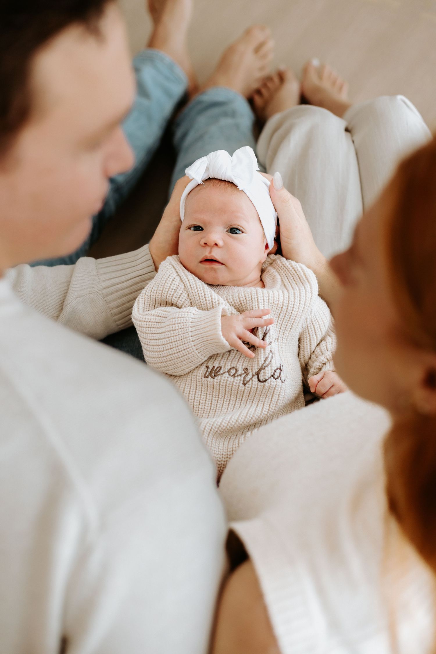 Newborn baby in beige knit romper with white bow headband held by parents in matching white sweaters.