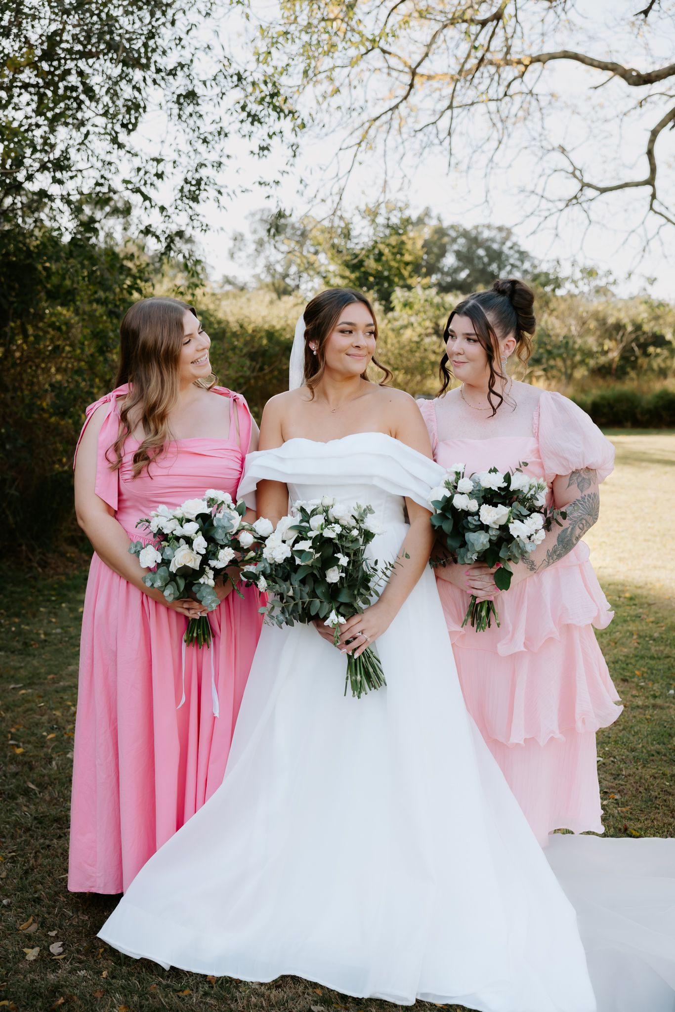 Bride in off-shoulder white gown poses with two bridesmaids in pink dresses holding white bouquets outdoors.