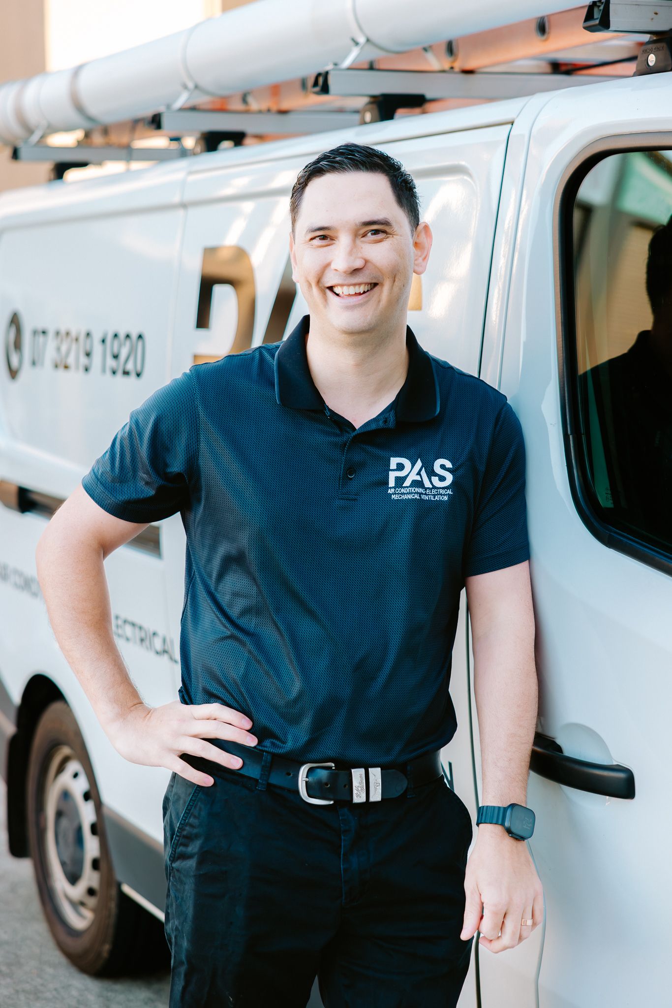 Smiling PAS electrician in navy polo shirt standing confidently beside a white company van with ladder rack.