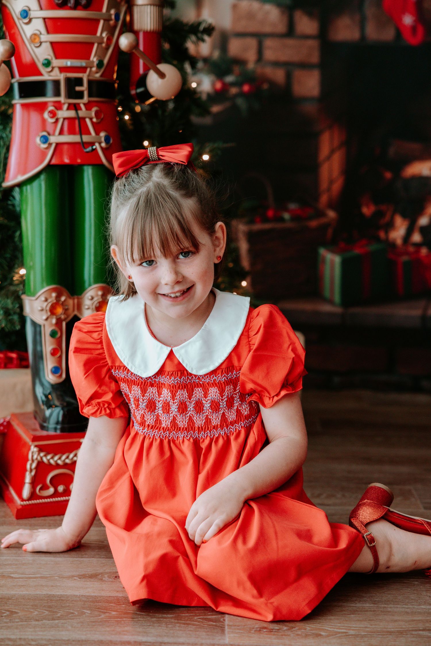 Young girl in red smocked dress with white collar sits on floor near Christmas tree and nutcracker decoration.