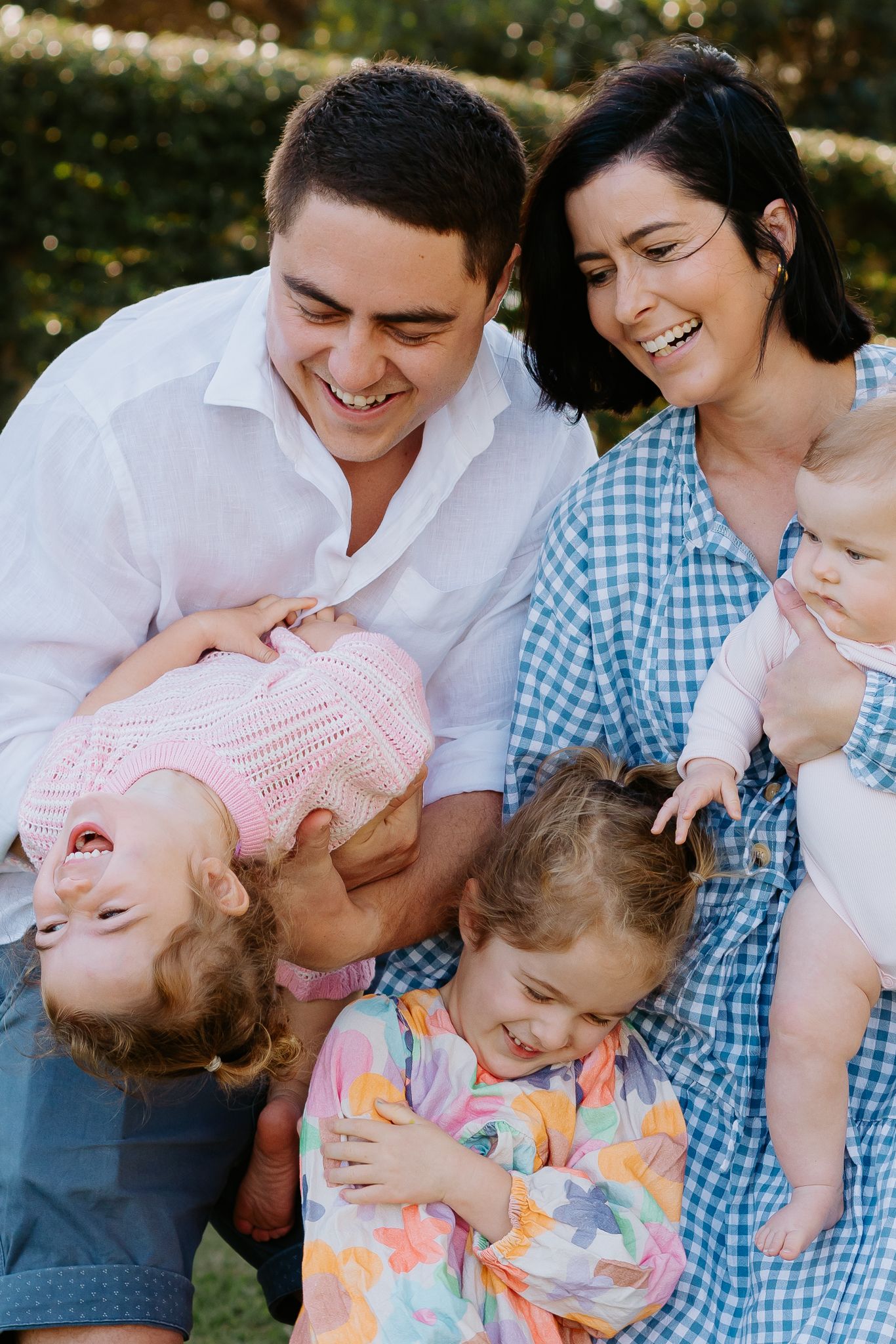 Happy family of five laughing outdoors, parents holding three young children in a sunny garden setting.