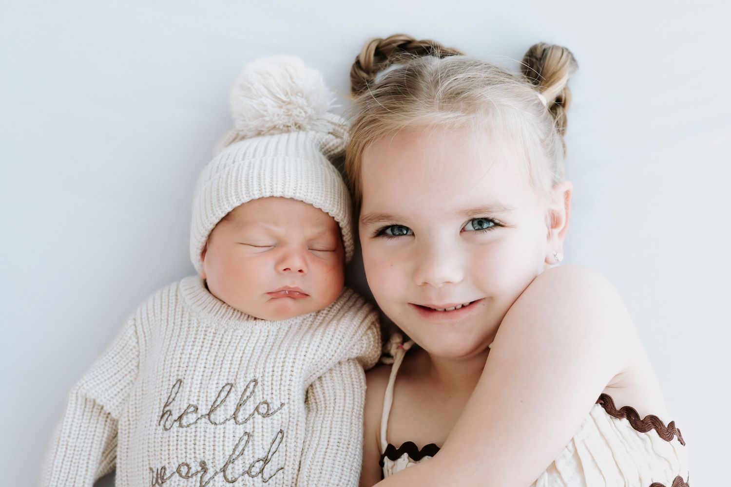 A smiling toddler girl with pigtails cuddles a sleeping newborn baby wearing a white hello world knit outfit.