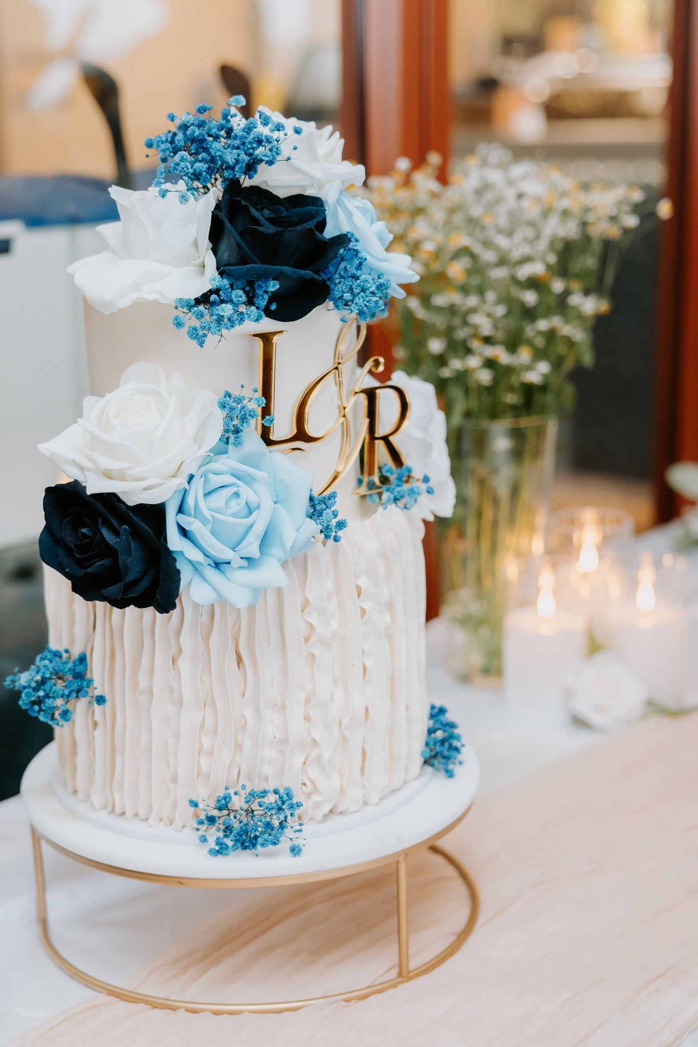 Elegant white wedding cake with blue and black roses, gold monogram initials, on a gold stand.