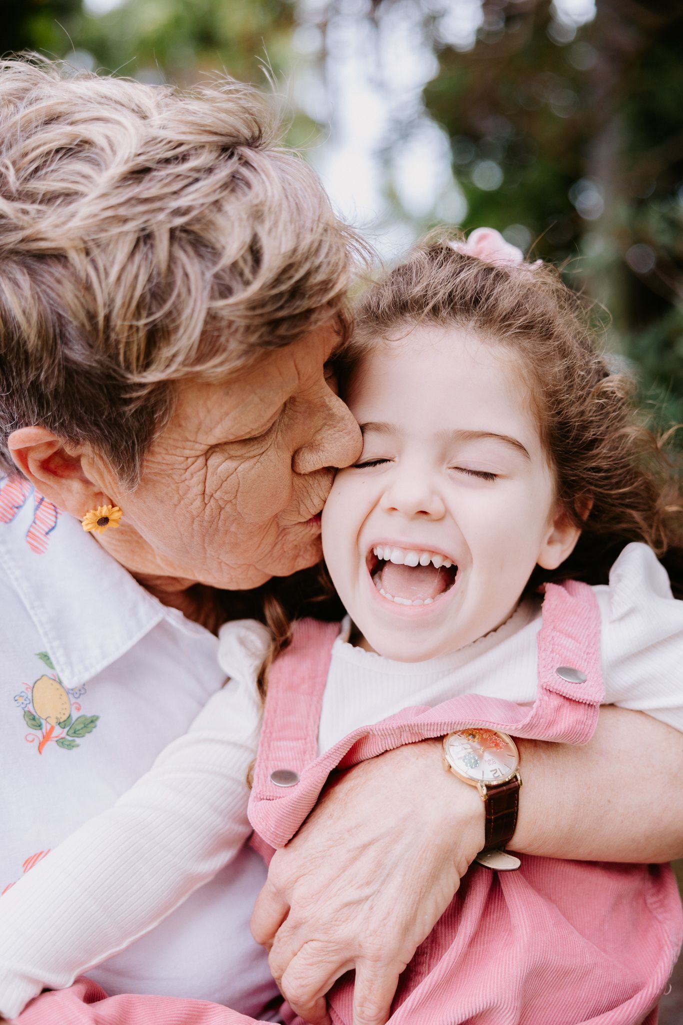 Elderly grandmother kissing laughing granddaughter on cheek outdoors, sharing a joyful moment together.