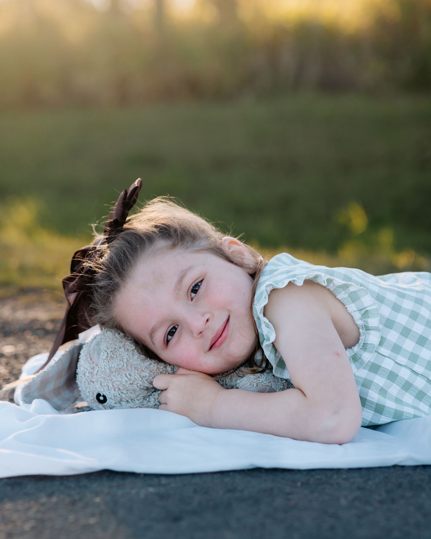 Smiling little girl in green gingham dress lying on white blanket outdoors, hugging a soft stuffed bunny toy.