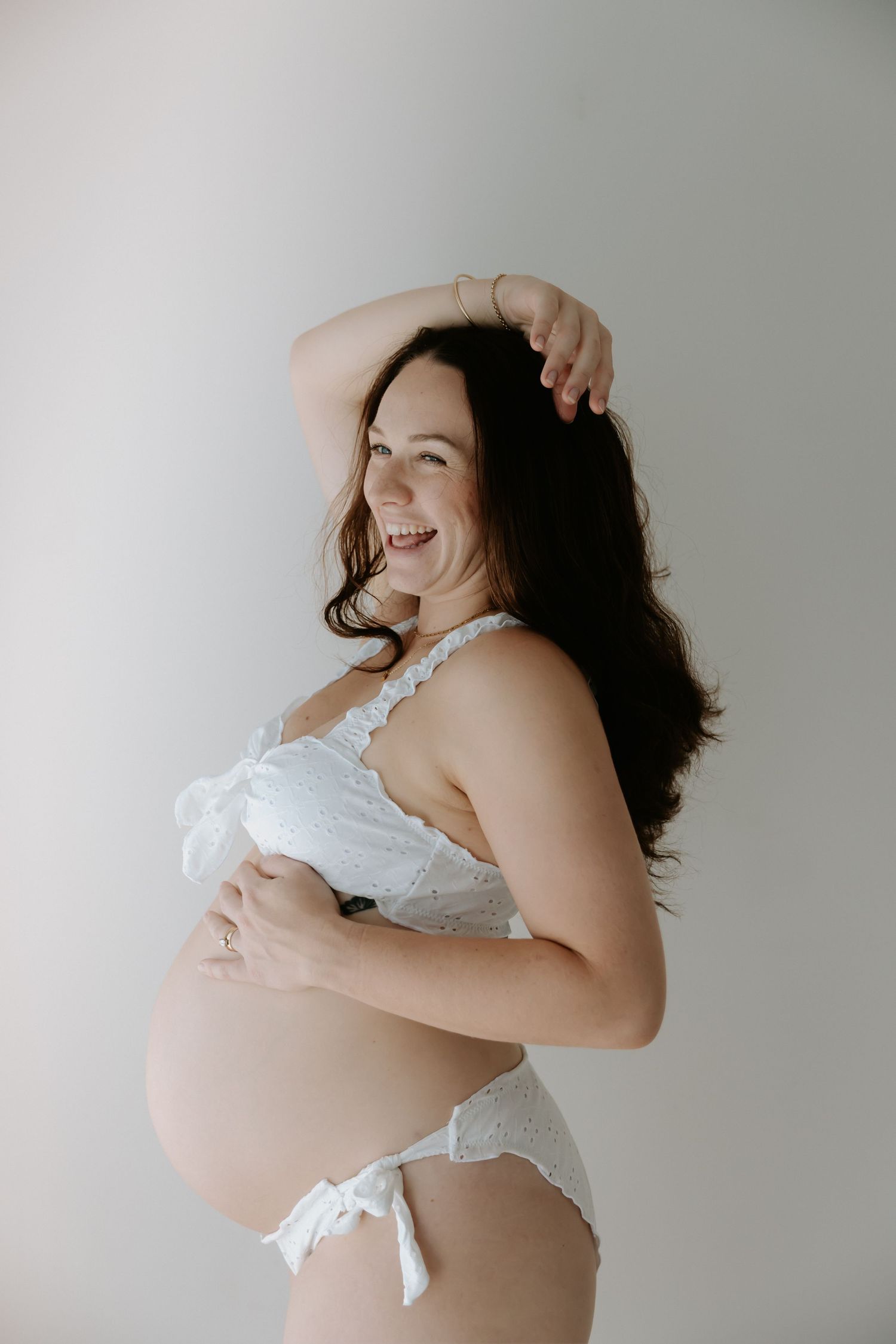 A joyful maternity photo series showing a woman in white lace embracing her pregnant belly against a neutral backdrop.