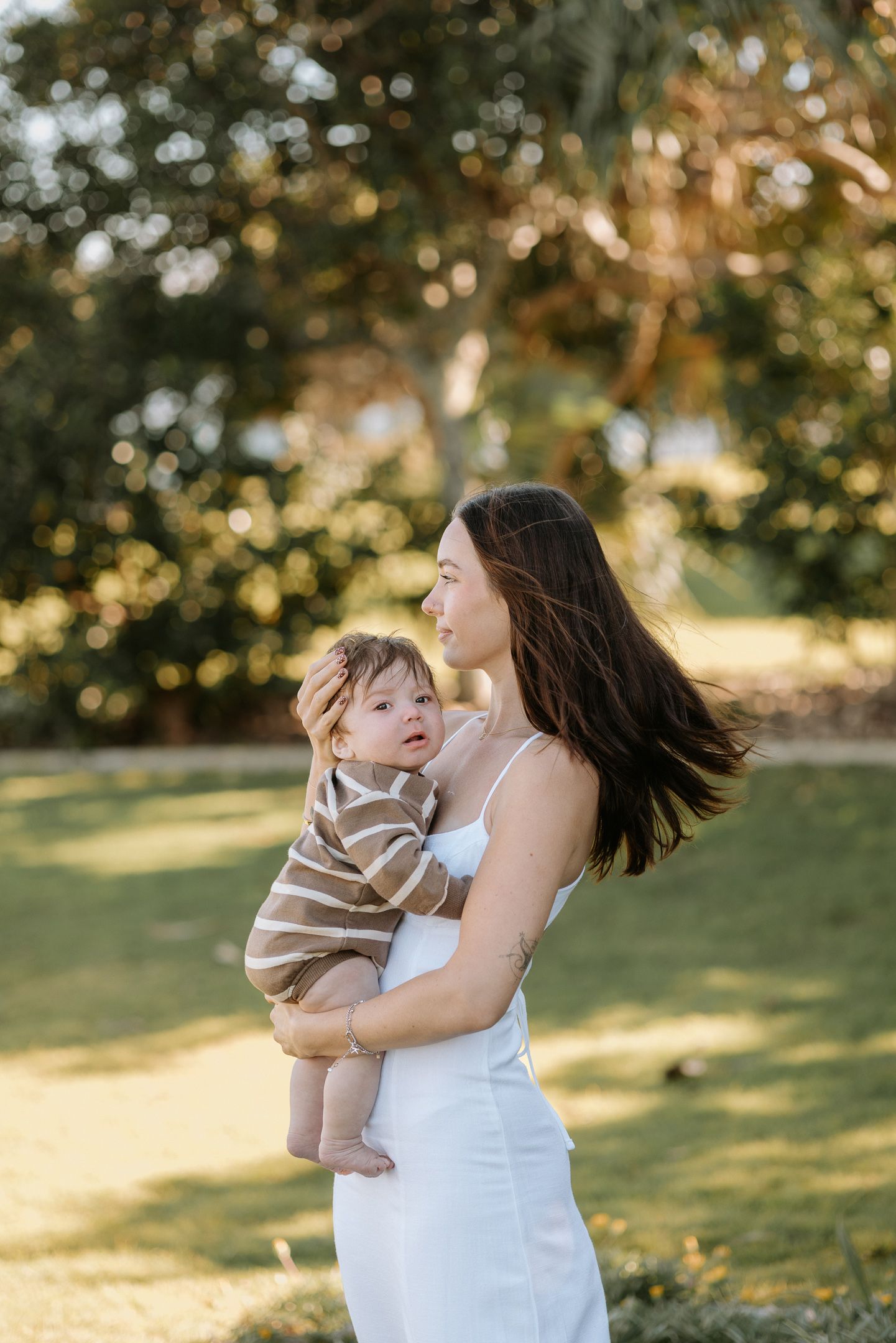 A woman in a white dress holds and plays with a small child in a park during golden hour sunlight.