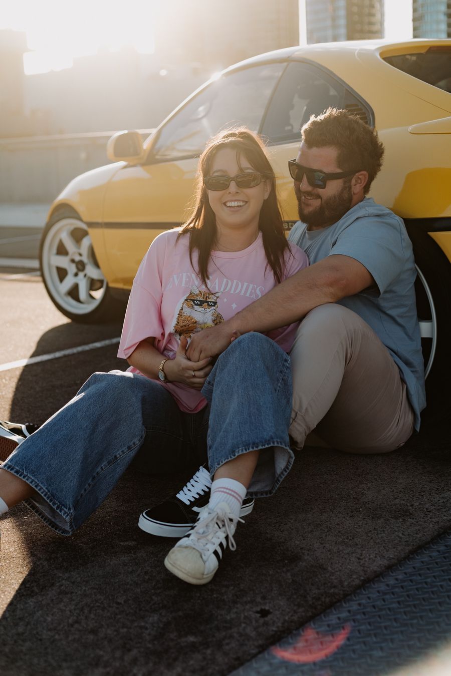 A couple sits together on the ground against a car in a sunlit parking lot wearing casual clothing.