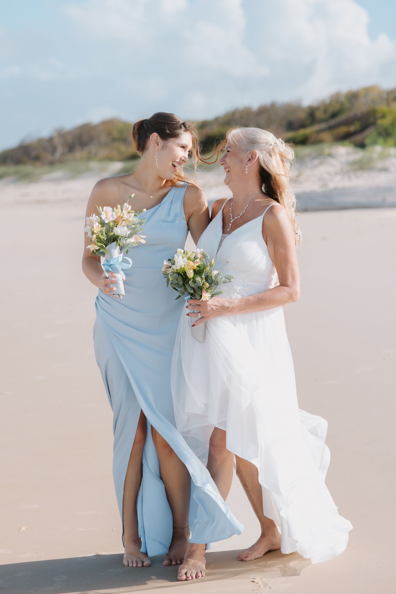 Two women in wedding attire walk barefoot on a beach, one in white bridal gown, other in blue bridesmaid dress.