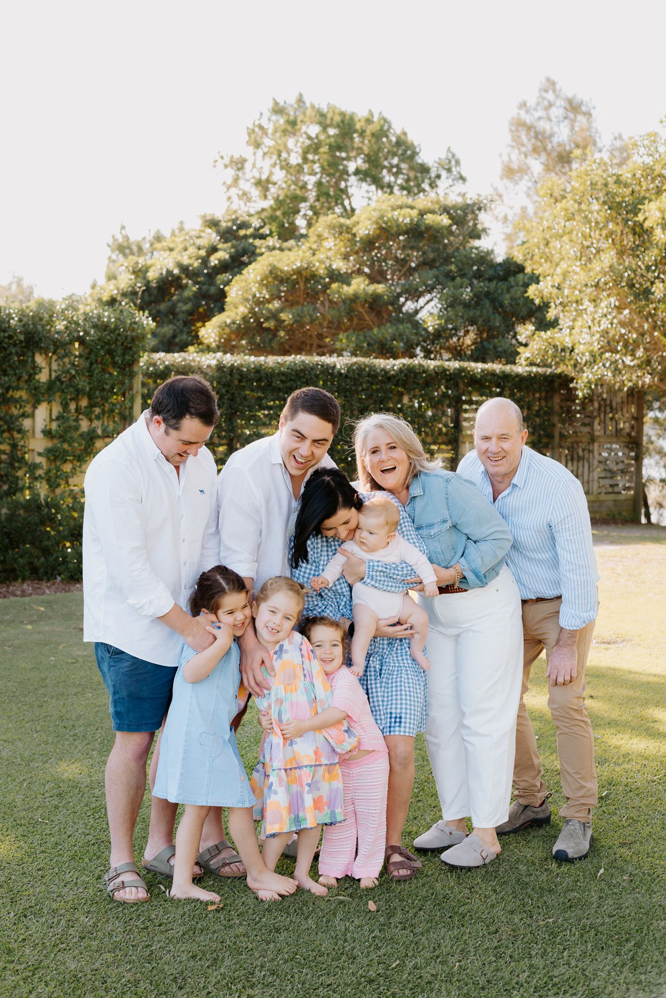 Large multigenerational family group smiling and laughing together outdoors on green lawn with lush hedge background.