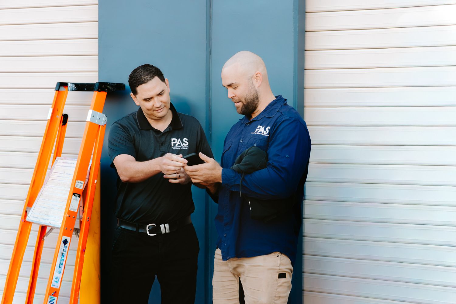 Two PAS technicians in branded uniforms reviewing work details beside an orange ladder outdoors.