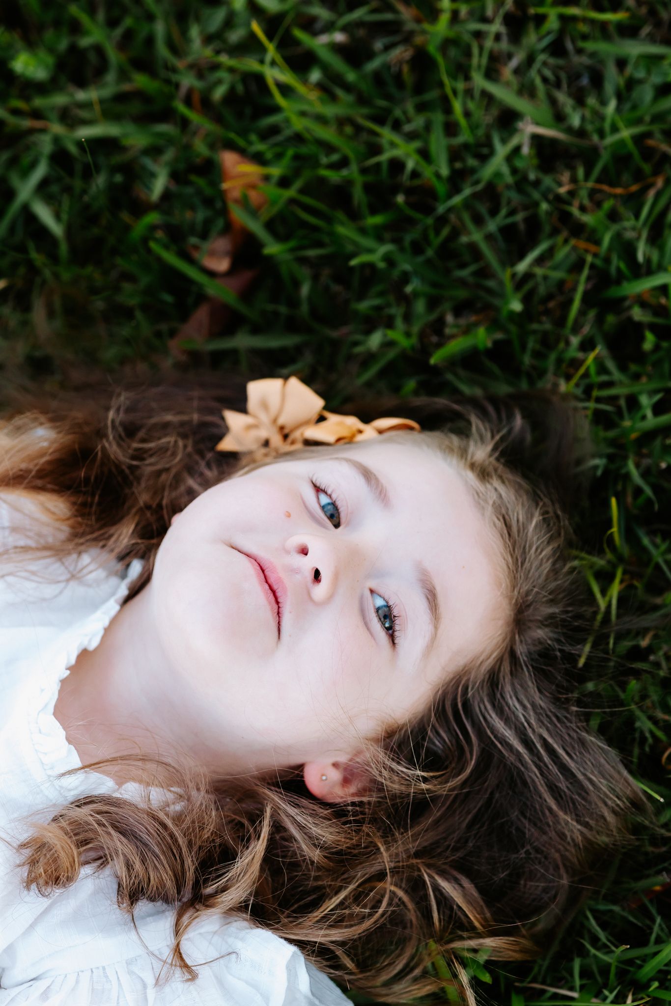 Young girl with curly hair lying on green grass, wearing white dress with autumn leaf, looking up peacefully.