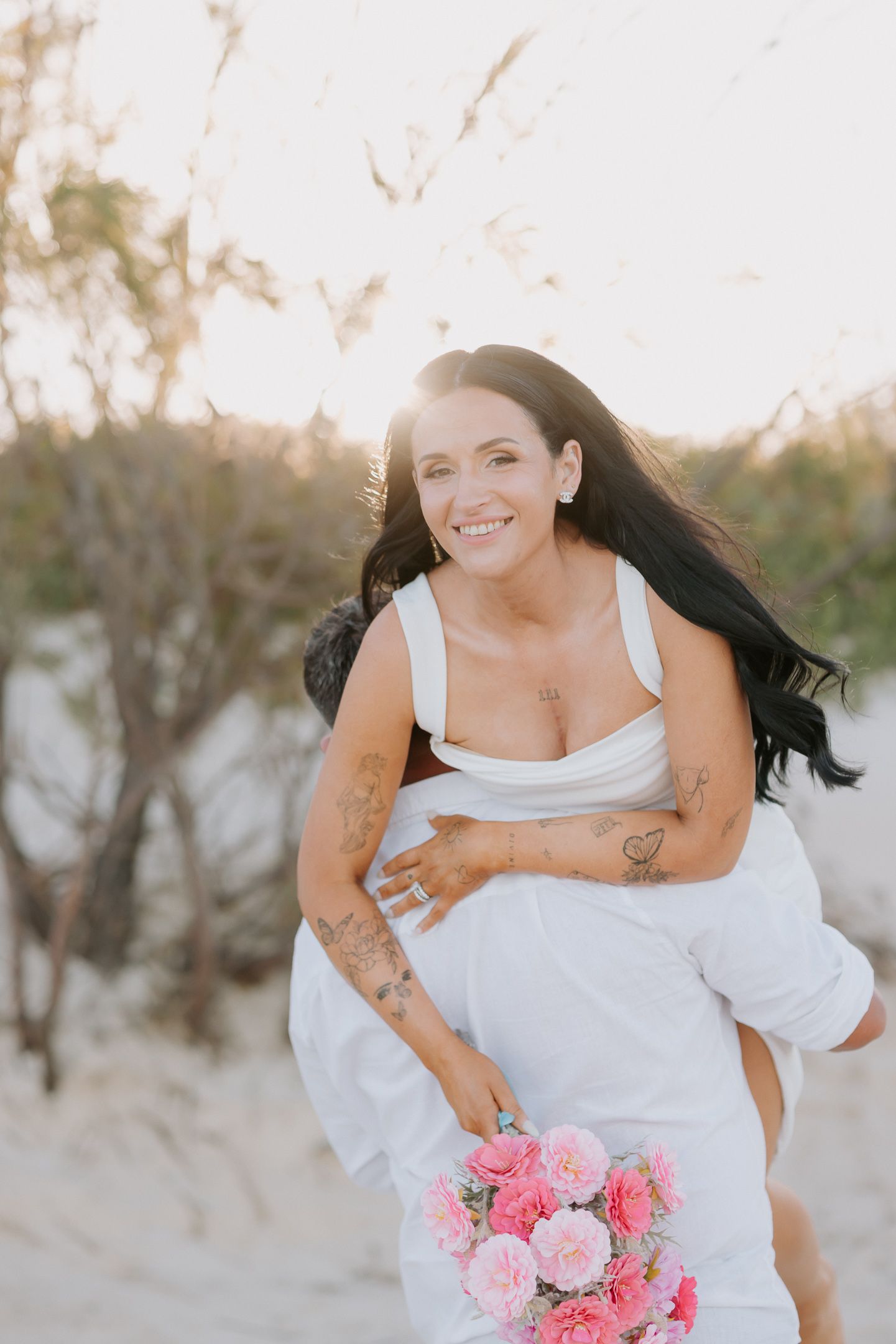 A woman in a white dress holds a pink bouquet while twirling joyfully on a beach at sunset.