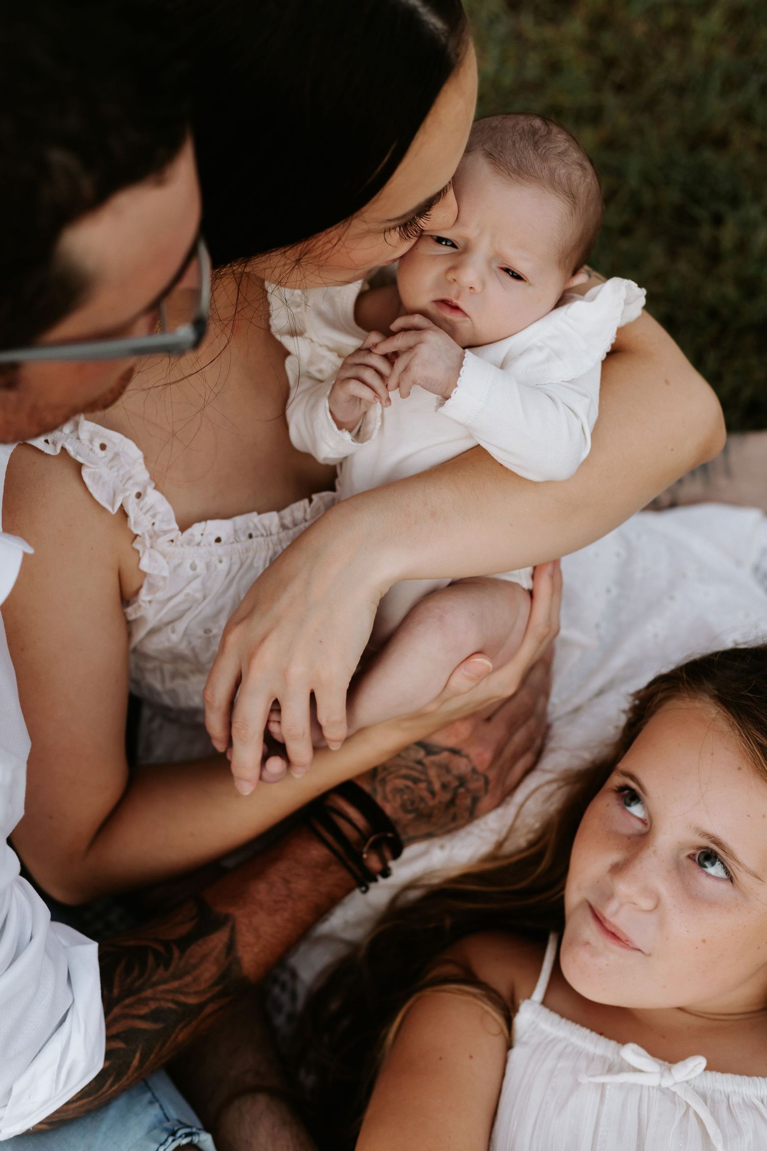 A tattooed couple sits outdoors holding a newborn baby in white, while a young girl in white dress looks up at them.