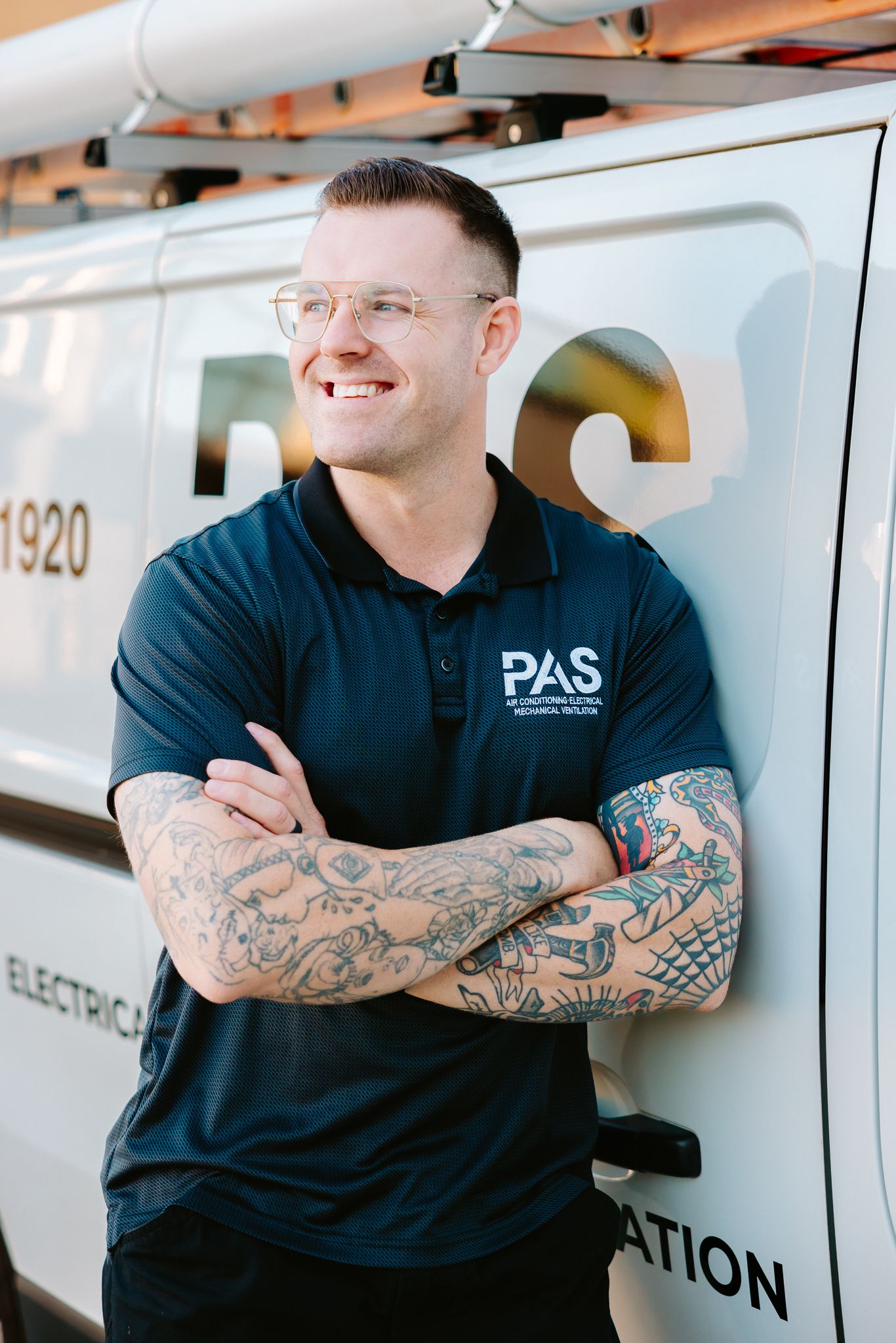 Smiling PAS technician with tattoos and glasses leans against a company van with arms crossed.