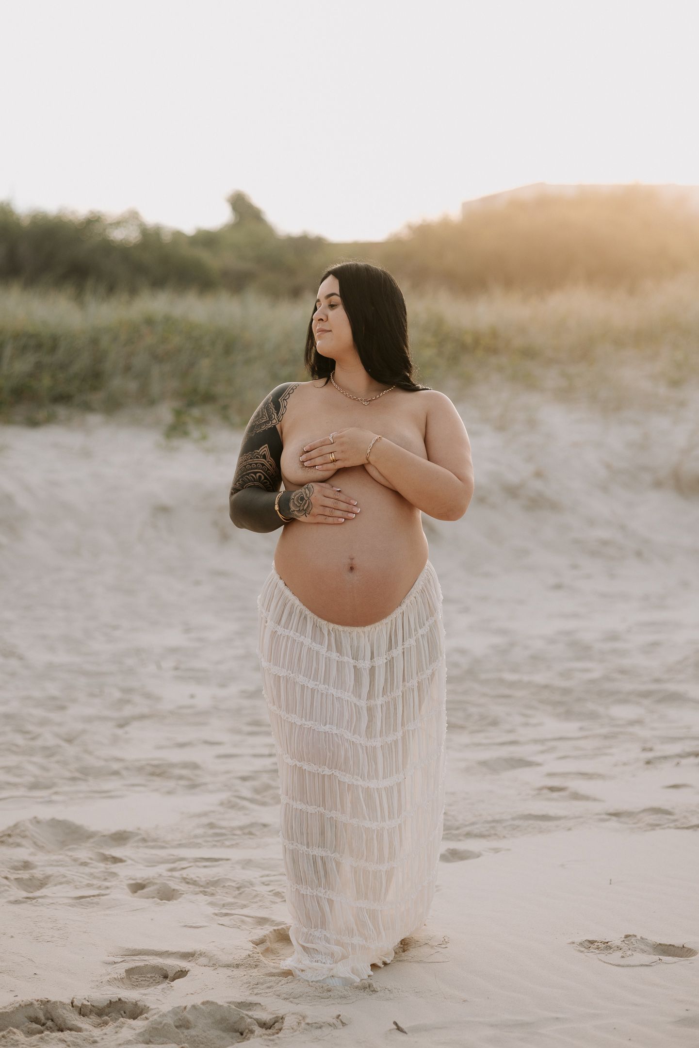Expectant mother in flowing white dress poses on sandy beach at sunset during maternity photoshoot.