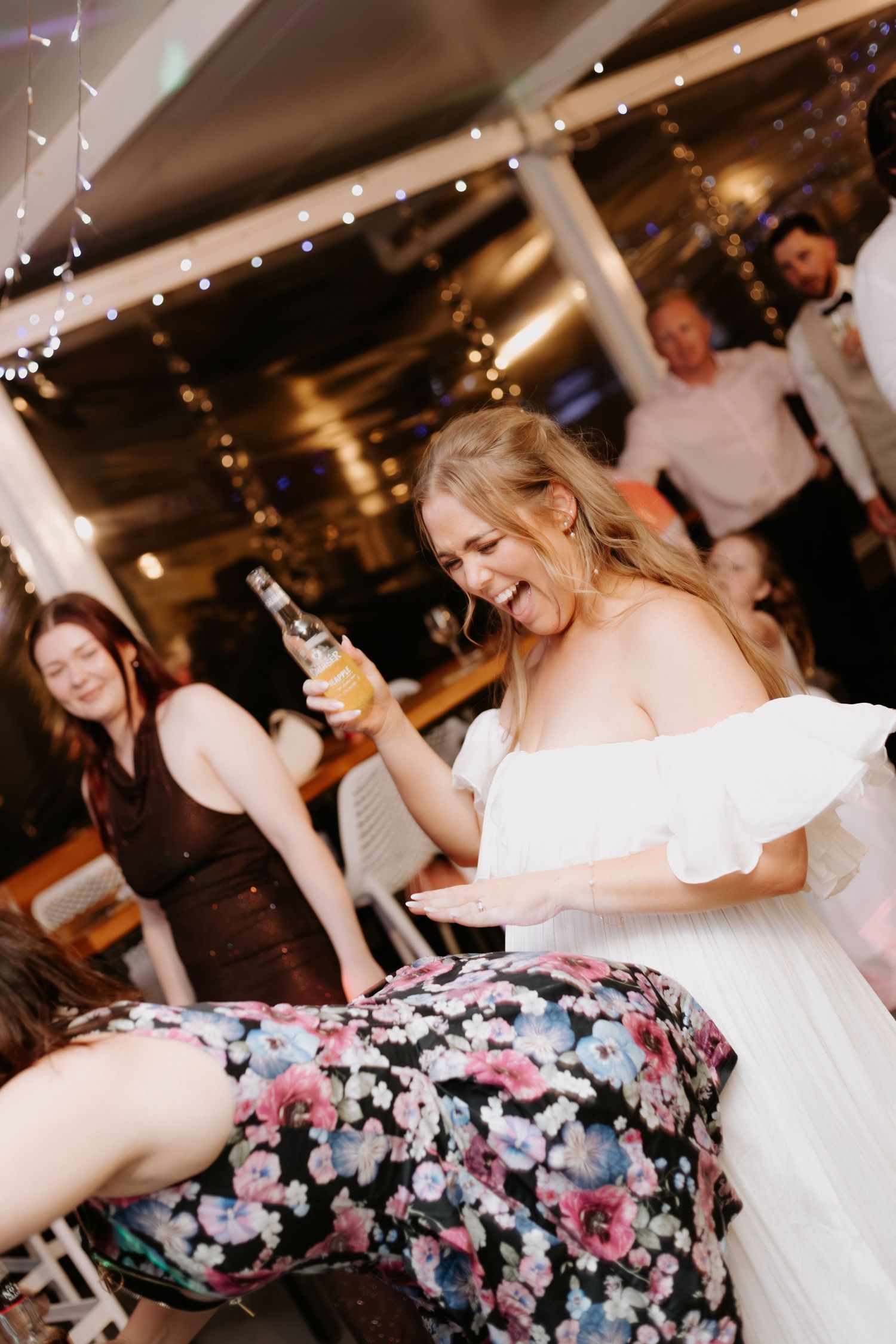 A group of people dancing and celebrating at what appears to be a wedding reception with twinkling lights in the background.