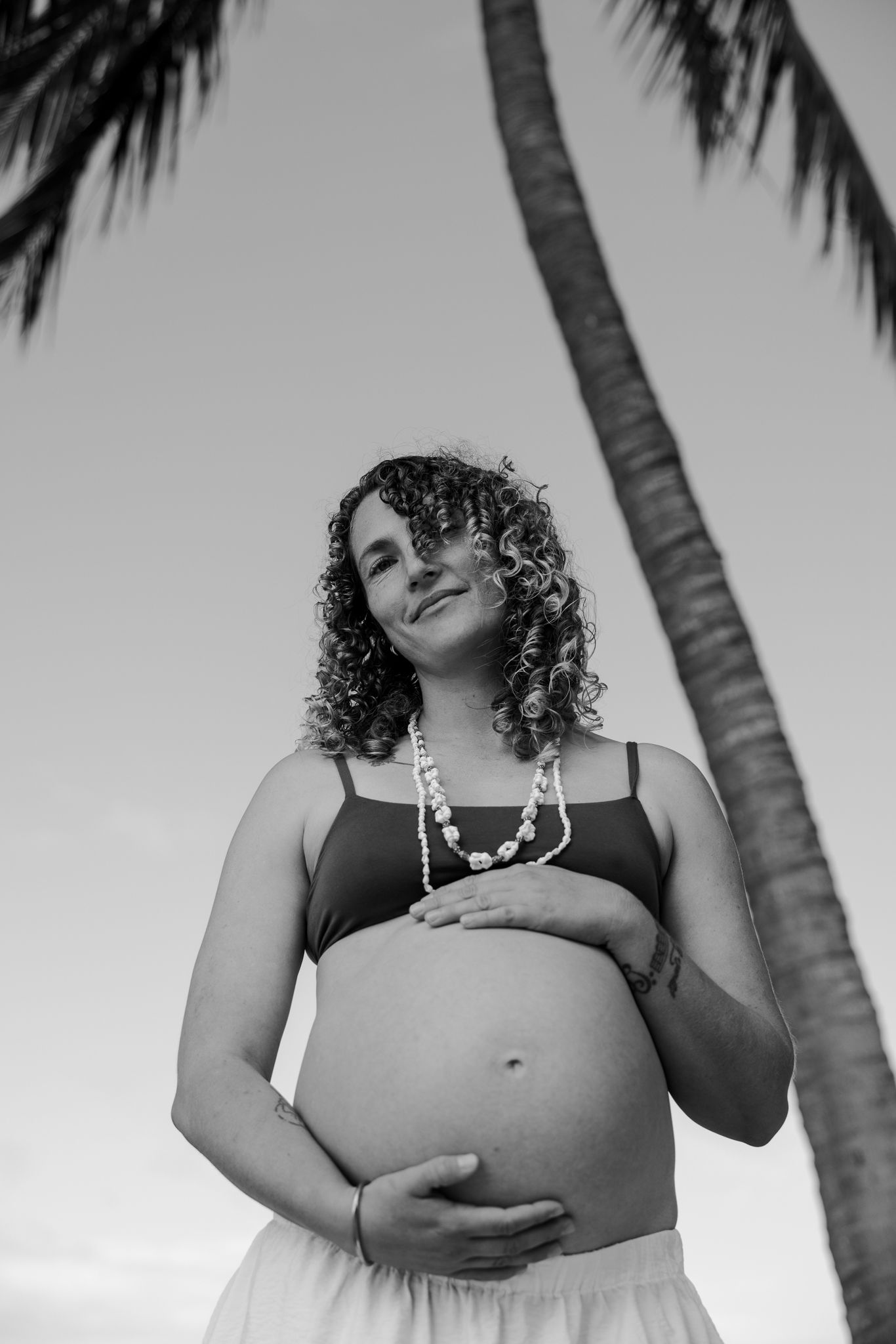 Black and white photo of a pregnant woman cradling her belly near a palm tree, wearing a beaded necklace.
