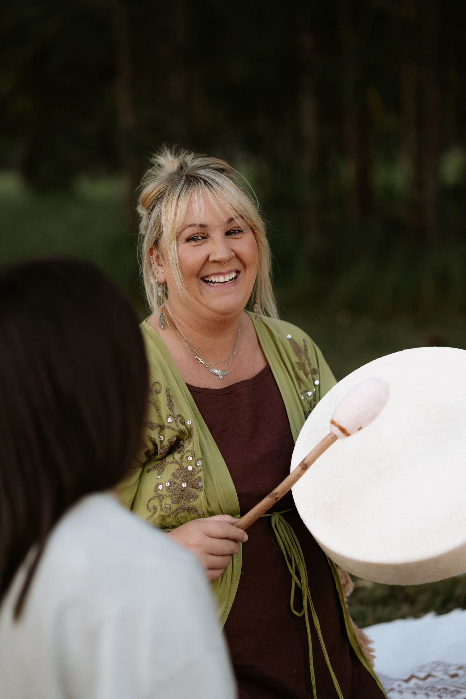 Smiling blonde woman playing a frame drum outdoors, wearing a green cardigan, in a lush forest setting.