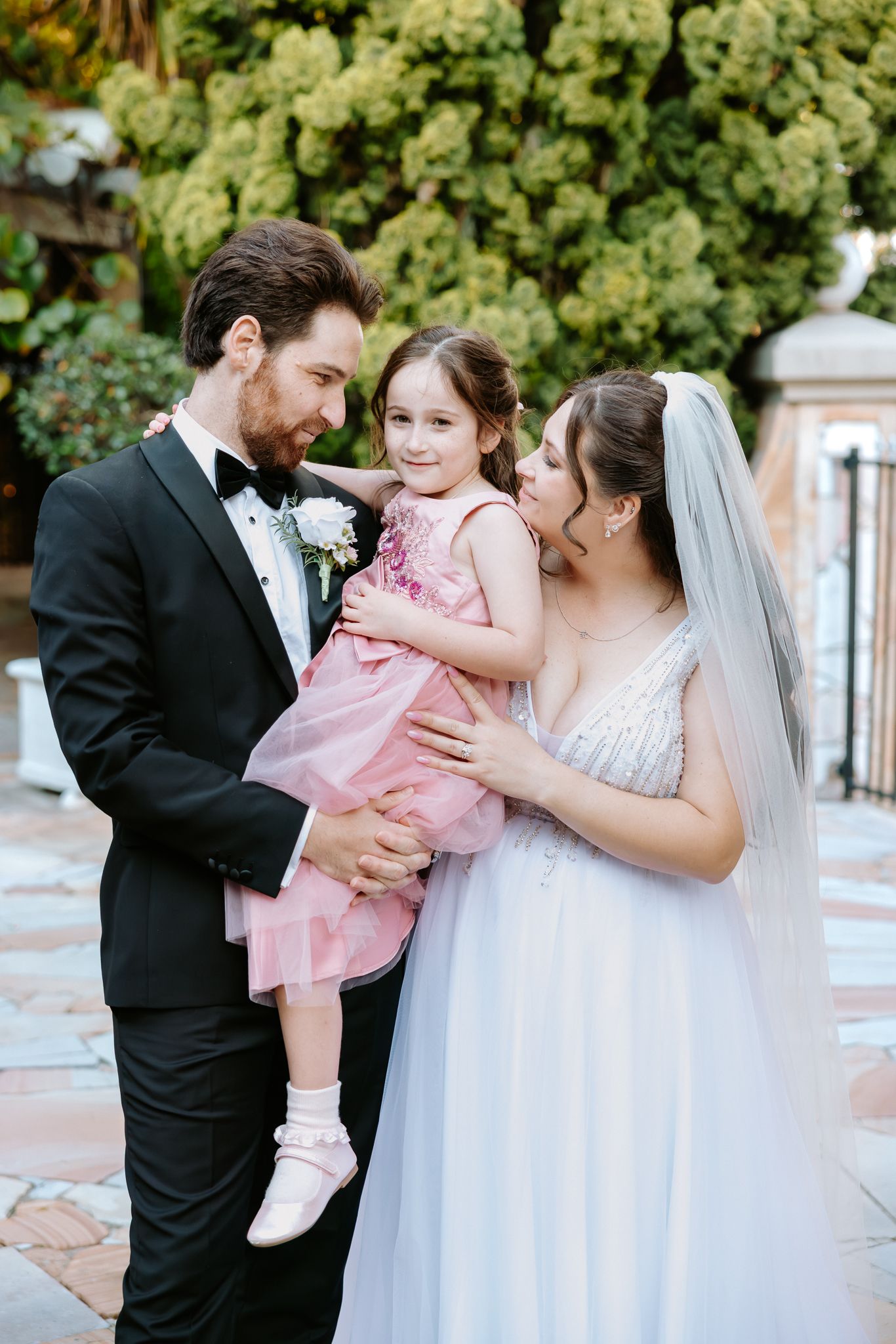 Bride and groom in wedding attire holding young girl in pink dress outdoors near lush green foliage.
