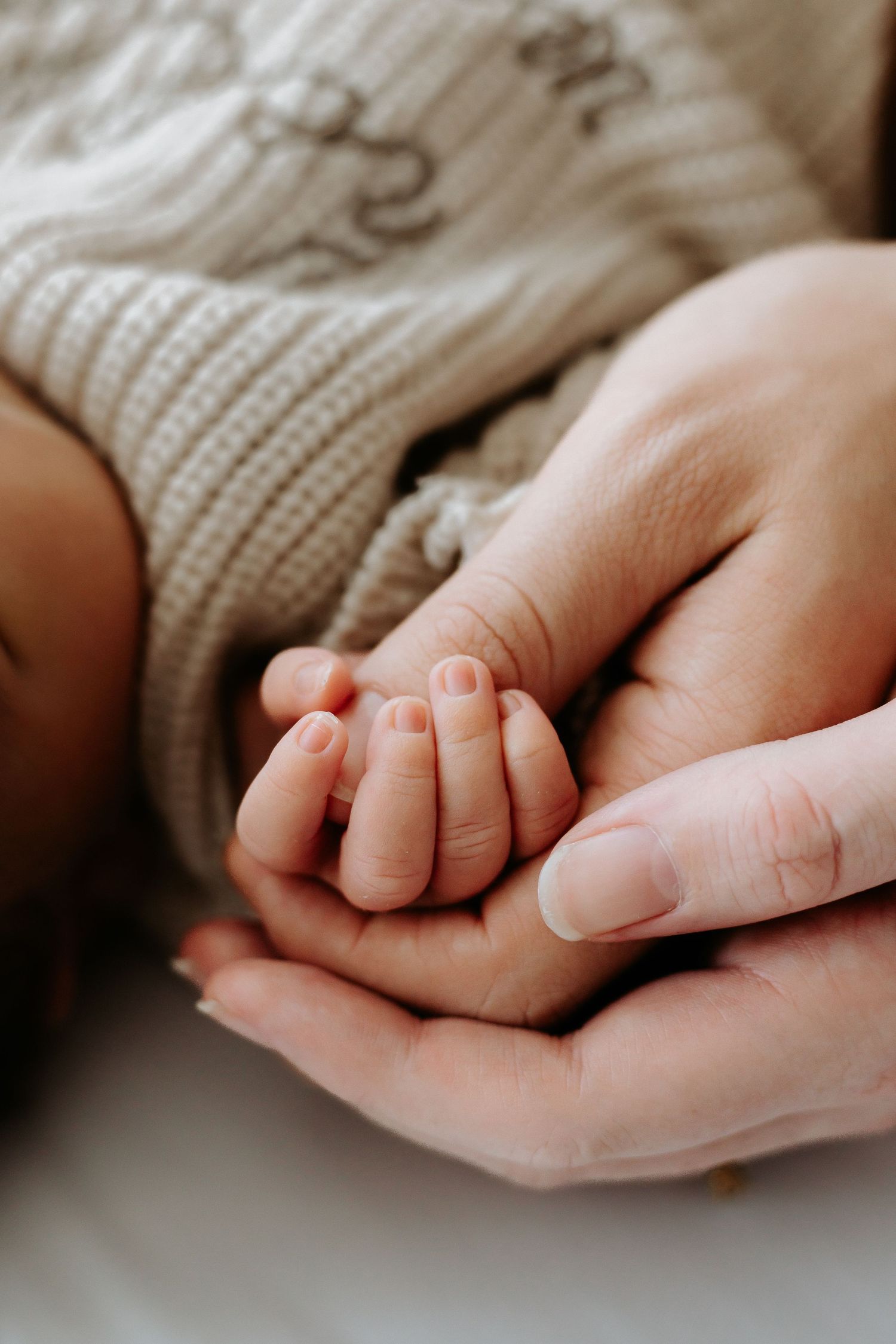 A parent's hands gently cradling a sleeping newborn baby's tiny fist, wearing a cozy knit sweater.