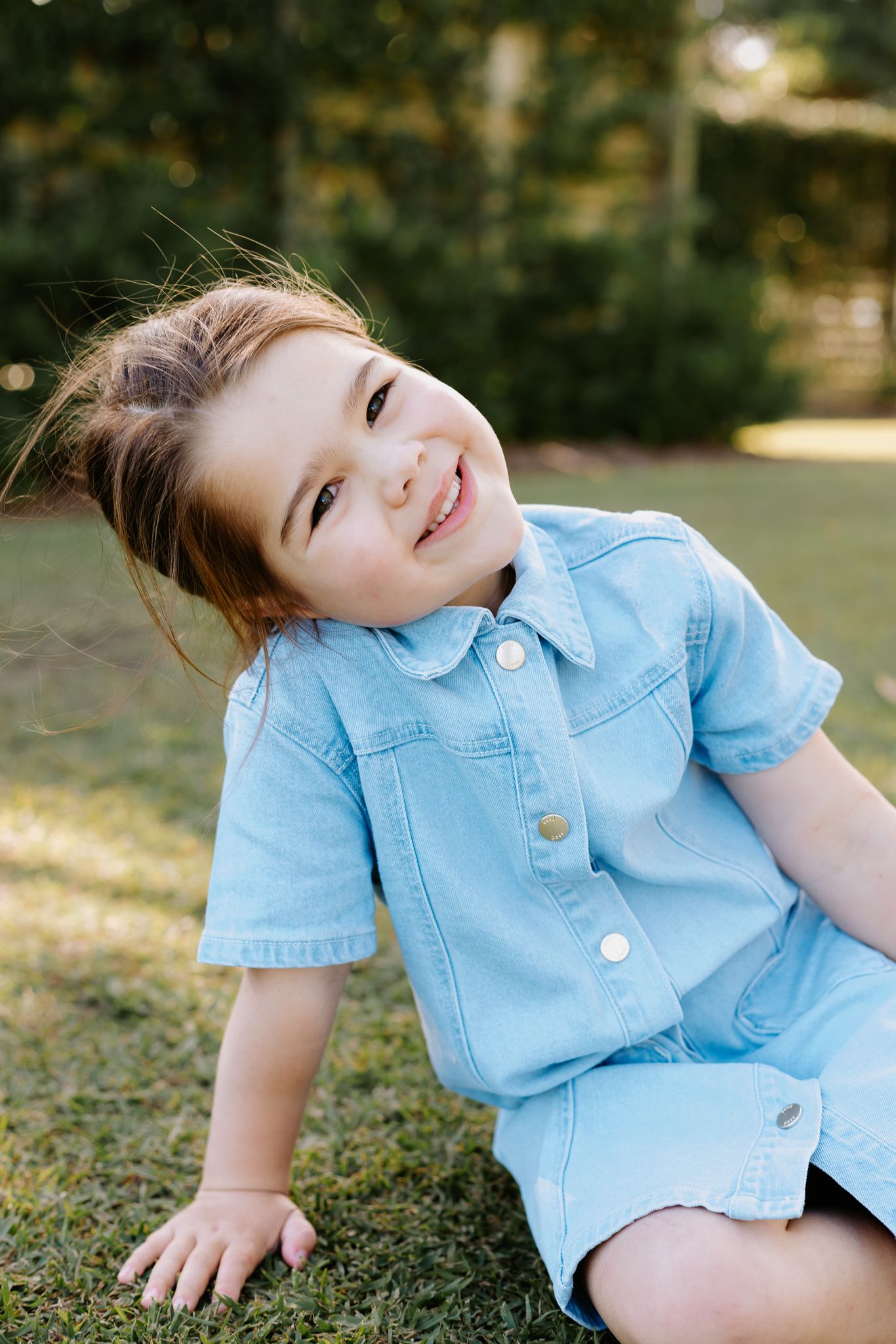 Young girl in blue denim romper sitting on grass, smiling playfully outdoors.
